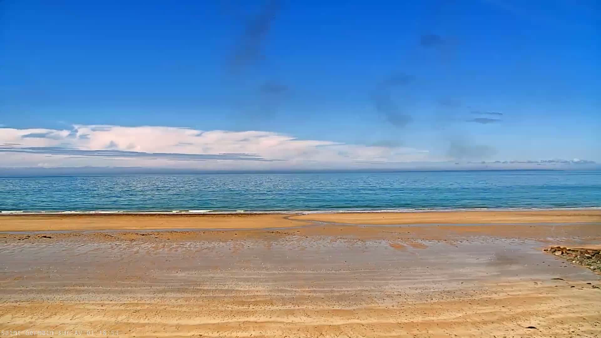 A wide sandy beach stretches to a calm, light blue ocean under a mostly clear blue sky with some fluffy white clouds.