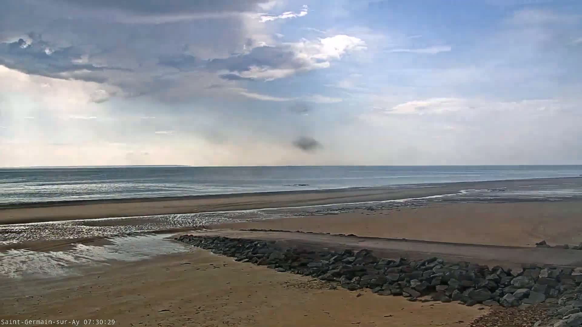 A sandy beach with a rocky embankment stretches toward a calm ocean under a partly cloudy sky.