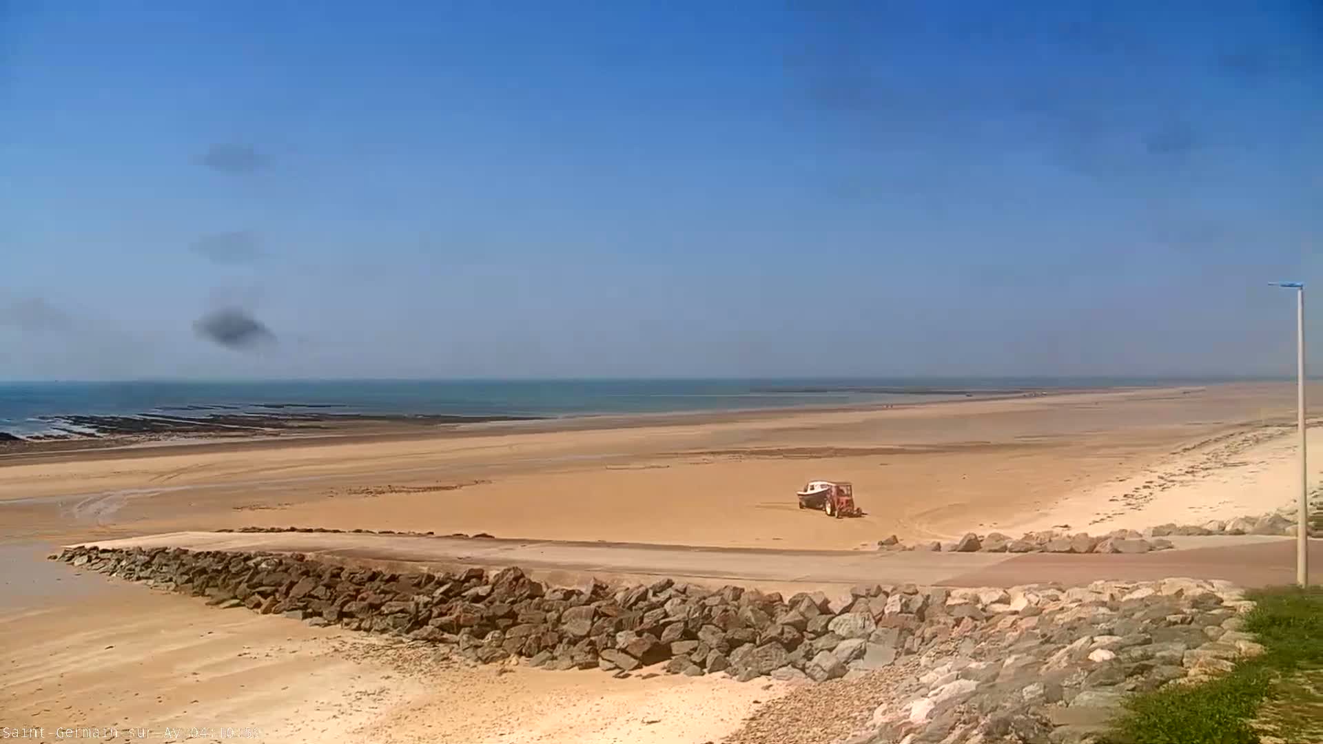 A sandy beach stretches to a calm ocean under a mostly clear blue sky with a few small clouds.