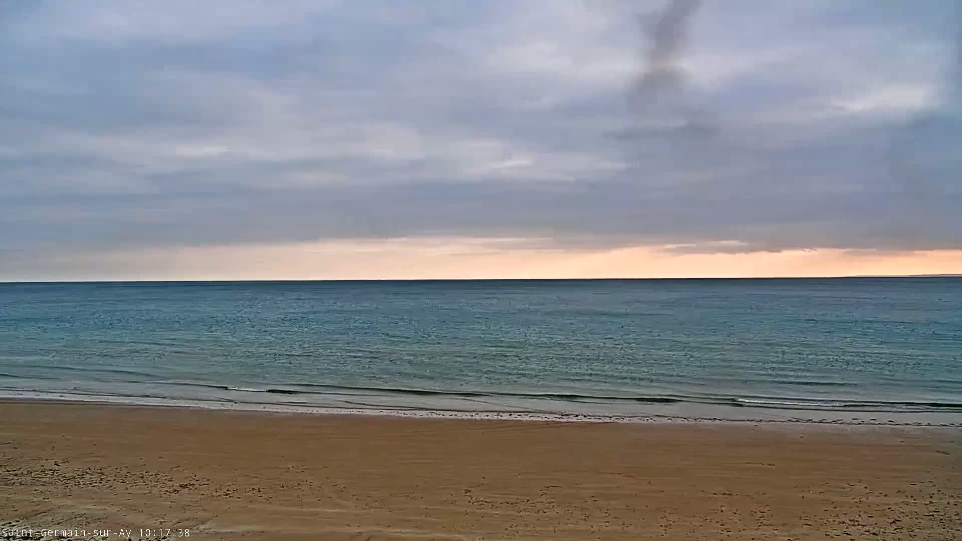 A sandy beach meets a calm, teal ocean under a mostly cloudy, pale sky.