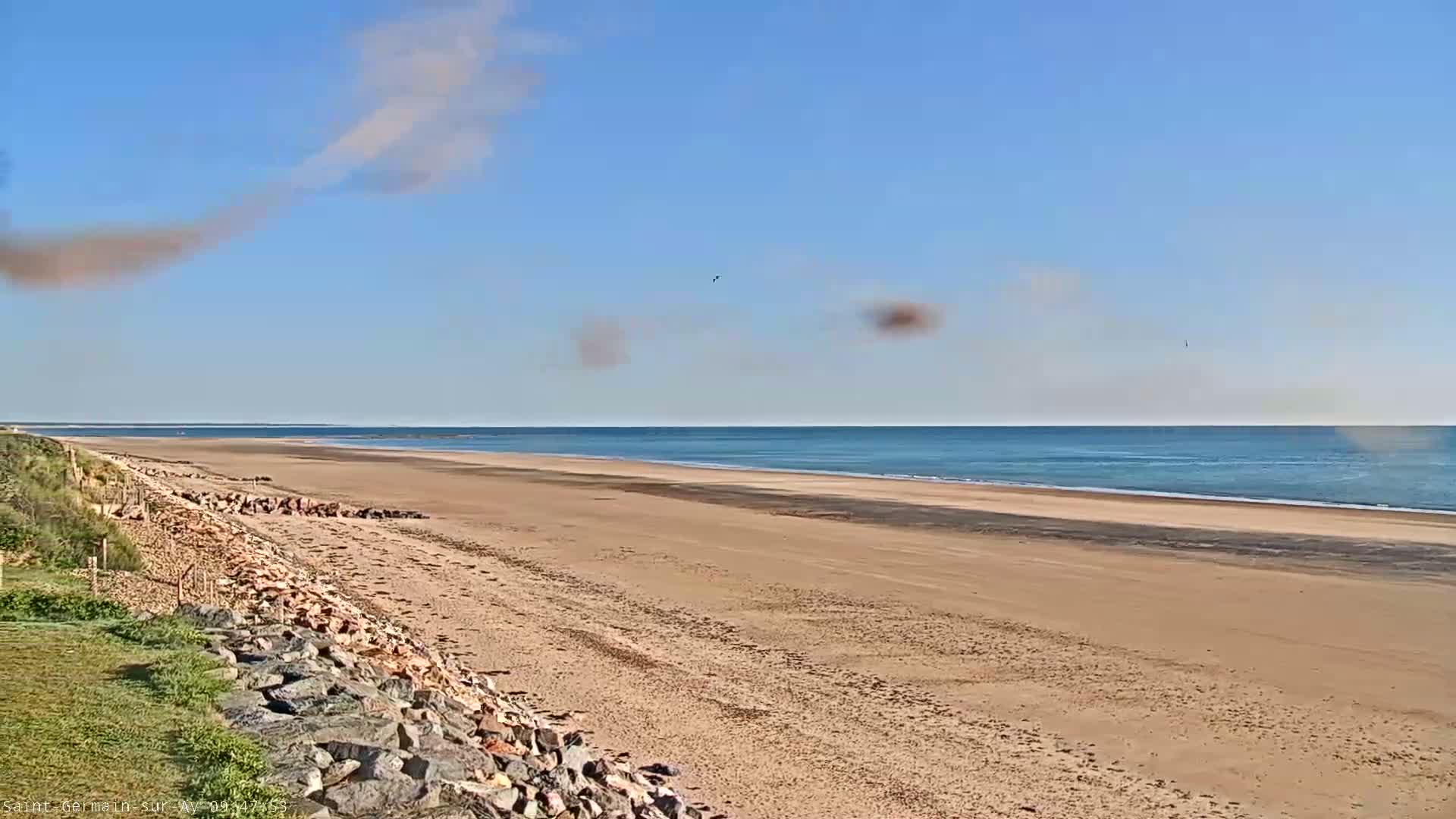 A sandy beach stretches to a calm, blue ocean under a mostly clear sky with a plume of light brown smoke or dust in the upper left corner.