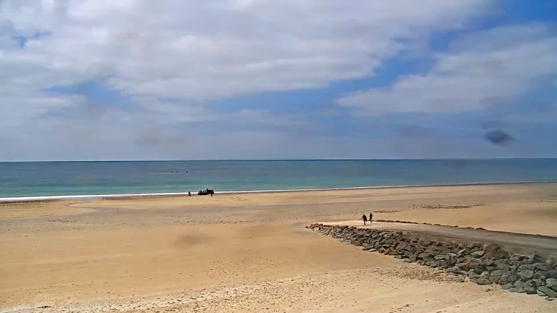 A sandy beach stretches to a calm ocean under a partly cloudy sky, with a few people and a vehicle visible on the shore.
