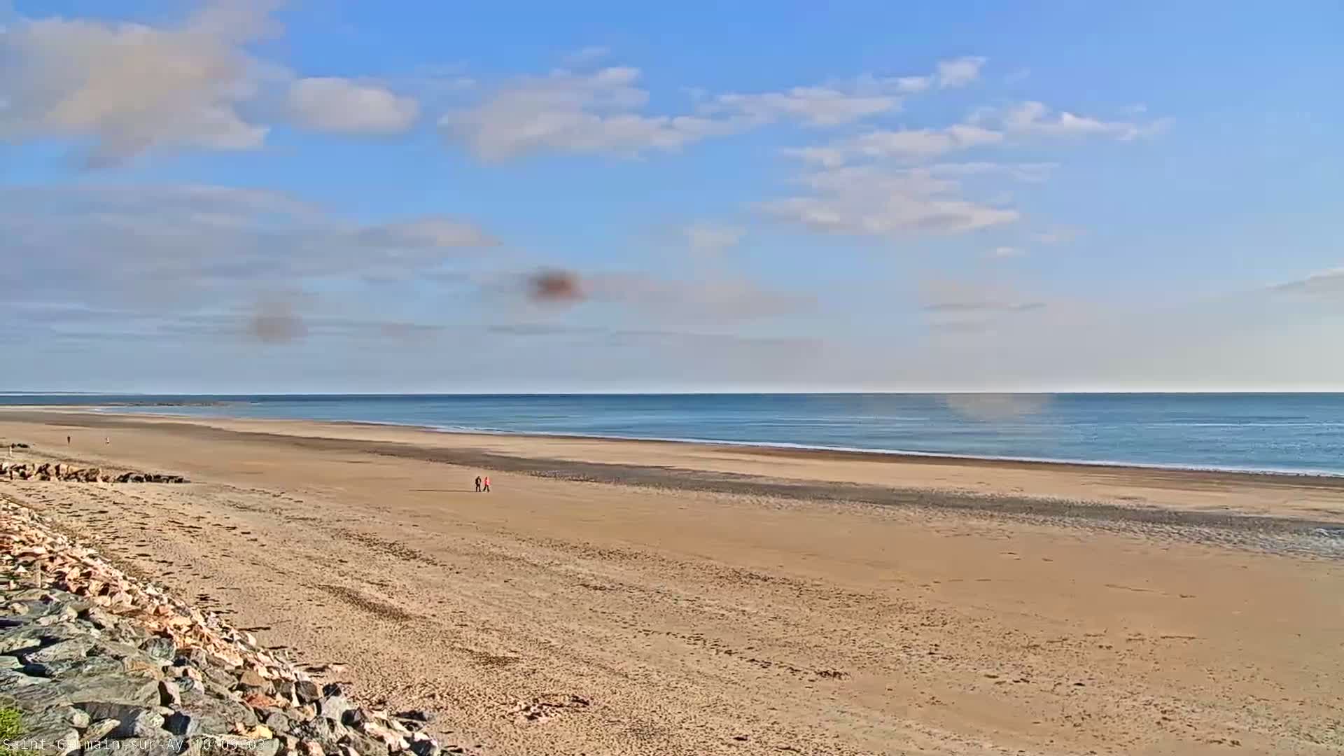 A long, sandy beach stretches to a calm, blue ocean under a mostly sunny sky with a few scattered clouds.
