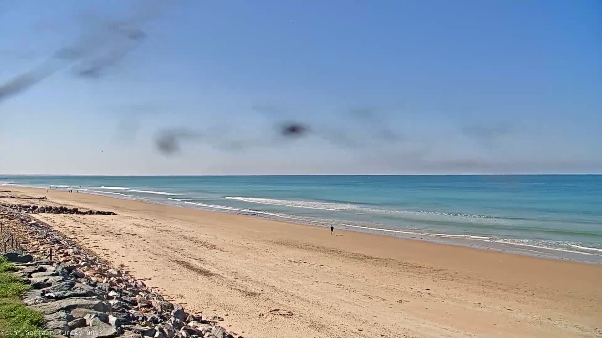 A long sandy beach stretches to a calm, light-blue ocean under a mostly clear sky with some dark smudges in the distance.