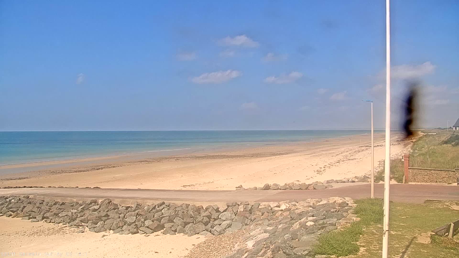 A wide sandy beach stretches to a calm ocean under a mostly sunny sky with a few scattered clouds.