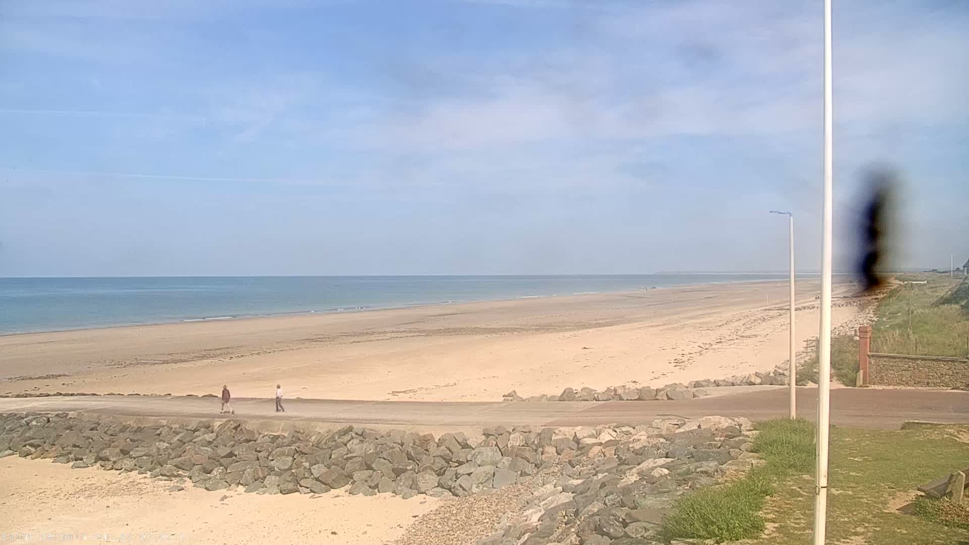 A wide sandy beach stretches to a calm ocean under a mostly clear, light blue sky, with two people walking on a paved path near the water's edge.