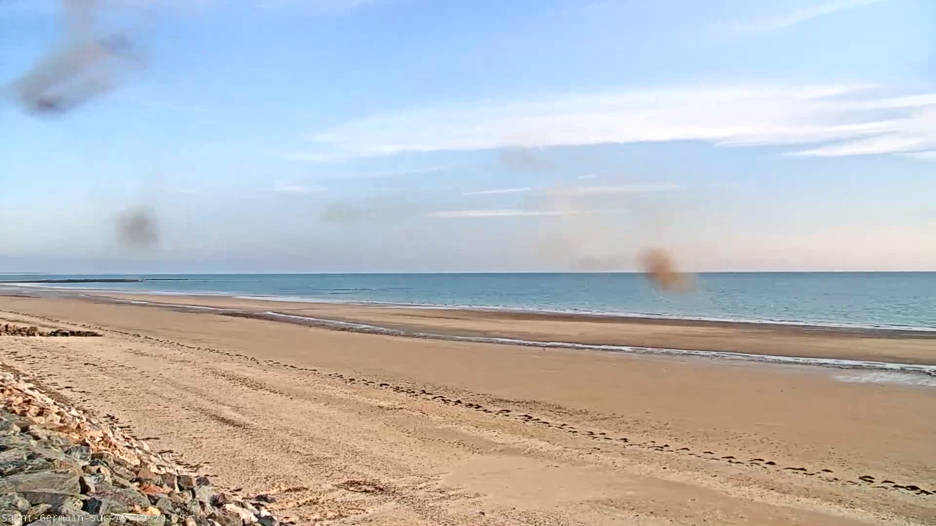 A wide sandy beach stretches to a calm ocean under a mostly clear, light blue sky with some scattered clouds and faint brown smudges.