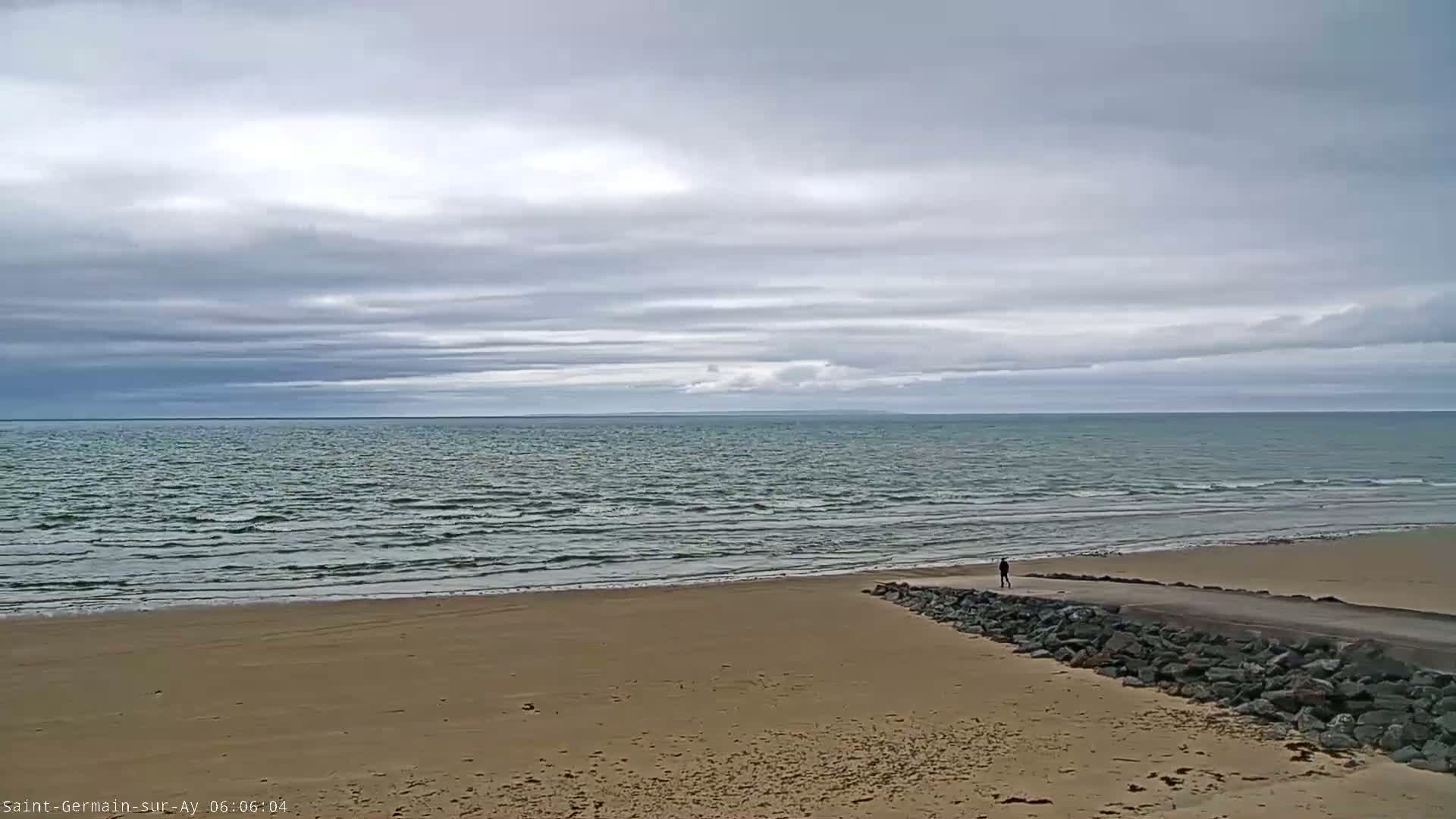 A person walks along a paved path that leads to a beach with gently lapping waves under an overcast sky.