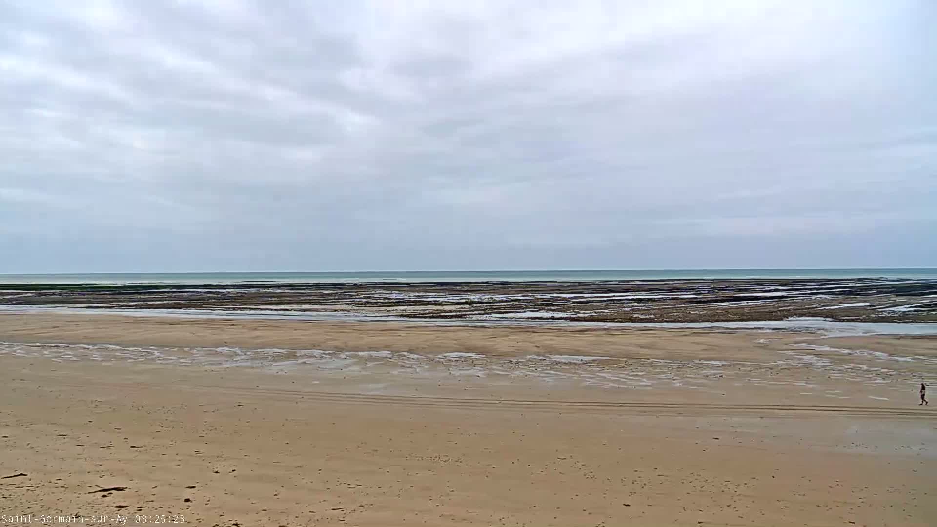 A person walks on a wide, sandy beach under an overcast sky toward a calm ocean with dark, intertidal rock formations.