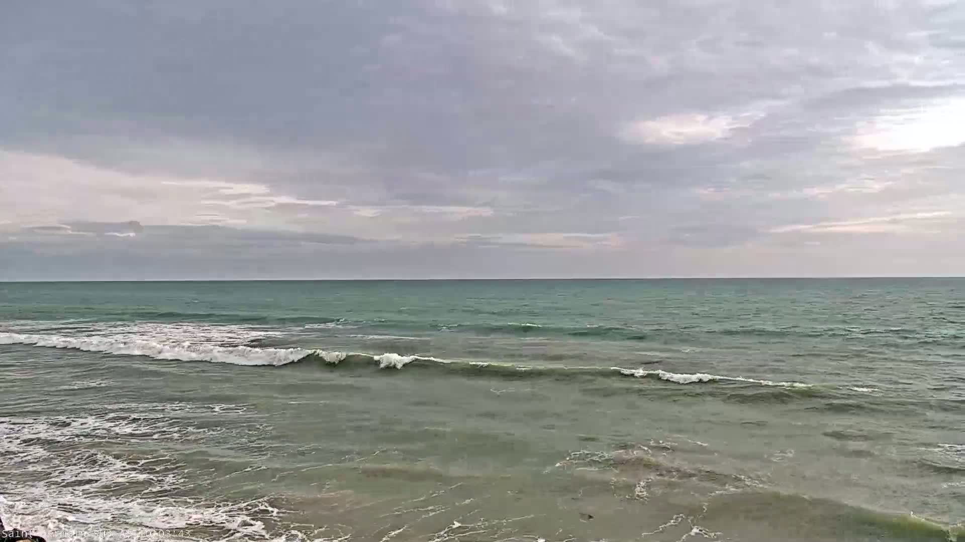 Under a cloudy sky, small waves roll onto a shallow, sandy shore.