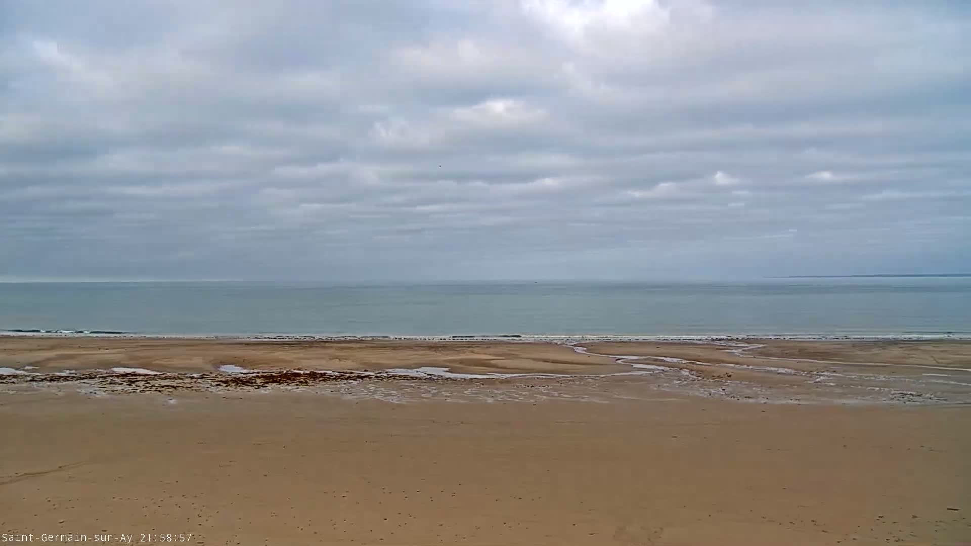 A mostly cloudy sky overlooks a wide, sandy beach and a calm, light-blue ocean.