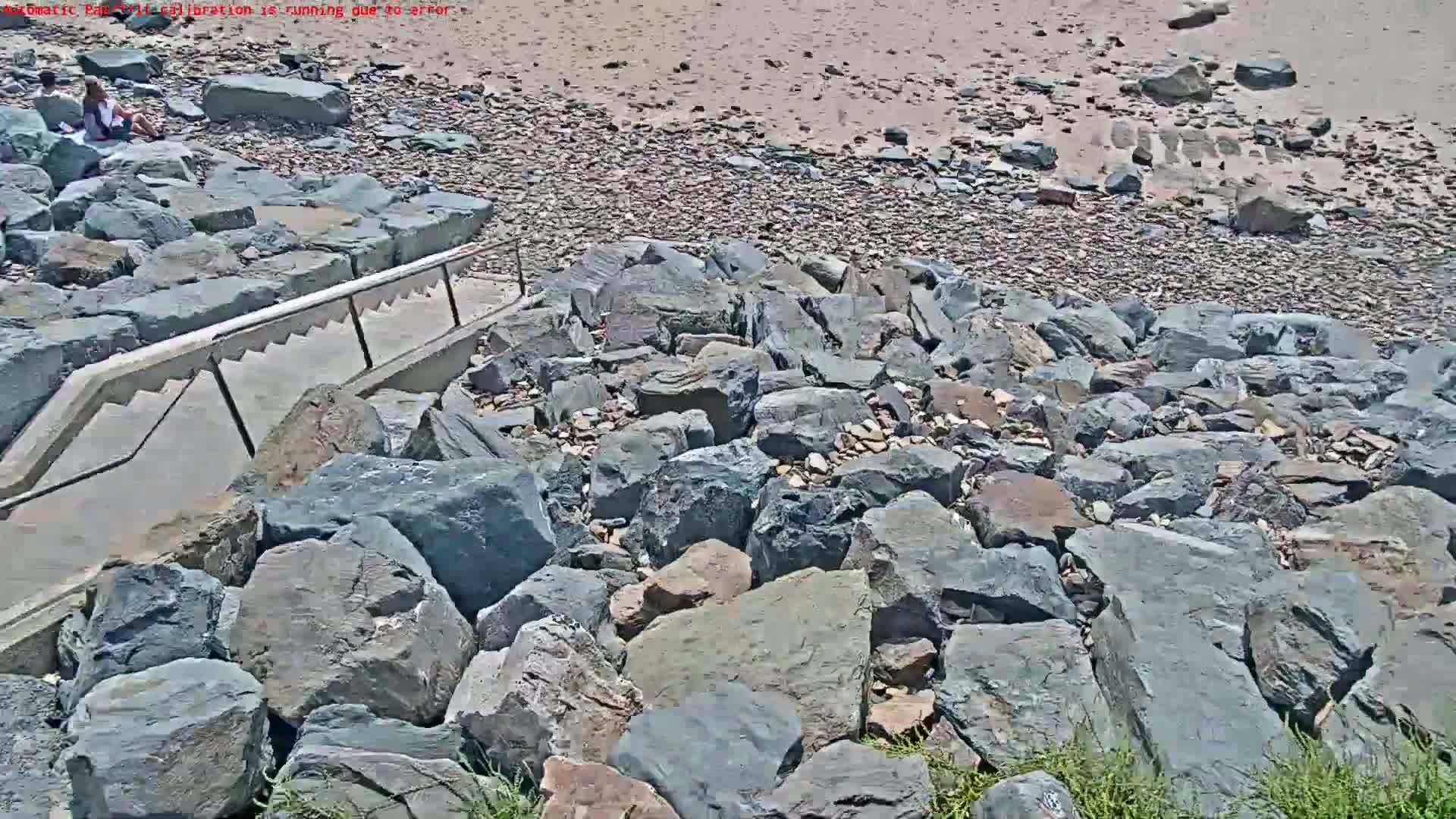 A concrete staircase with a metal railing descends to a rocky shoreline on a sunny day.