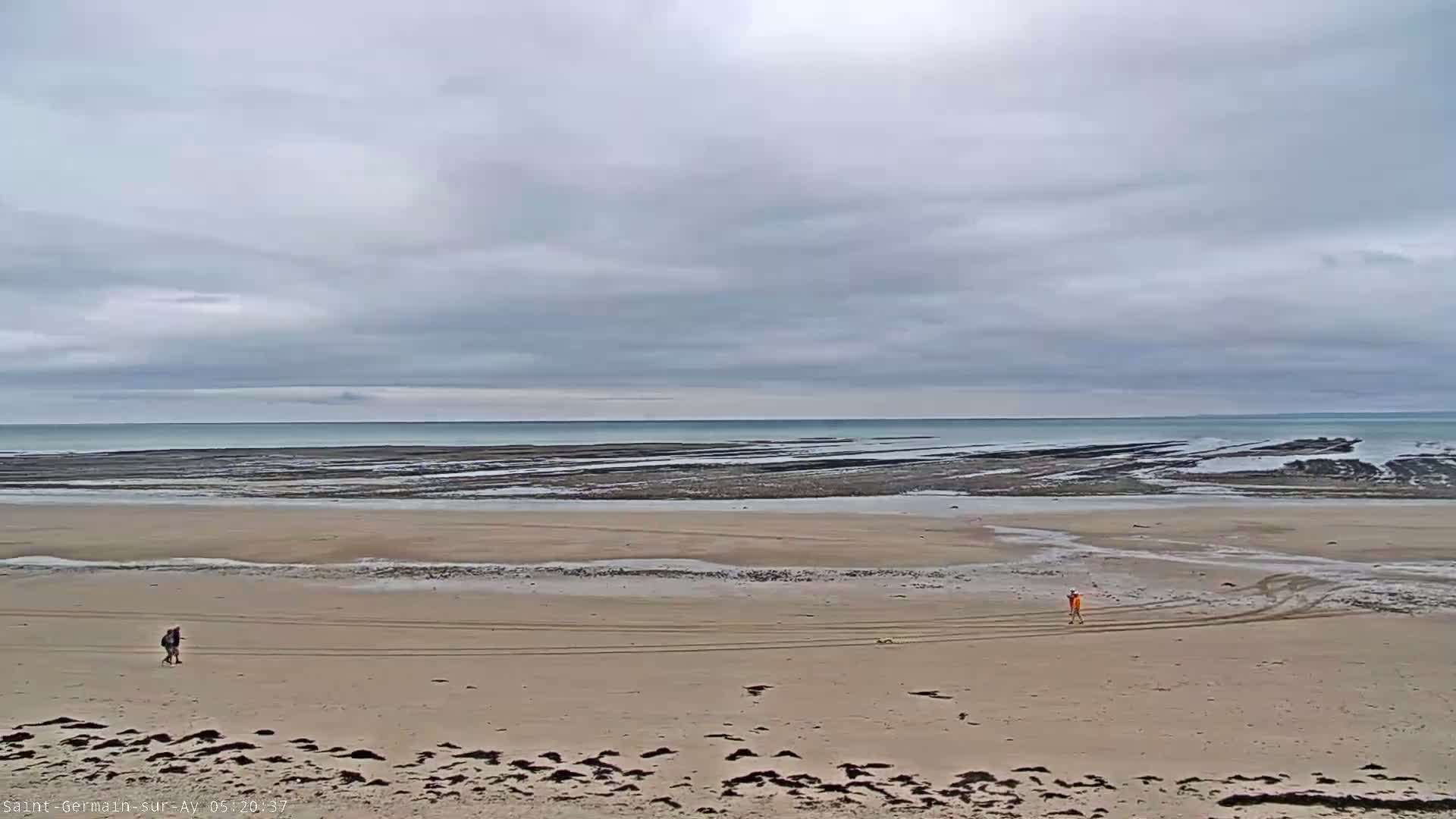 Two people walk on a wide, sandy beach under a cloudy sky, with a calm ocean visible in the distance revealing dark, textured intertidal zones.