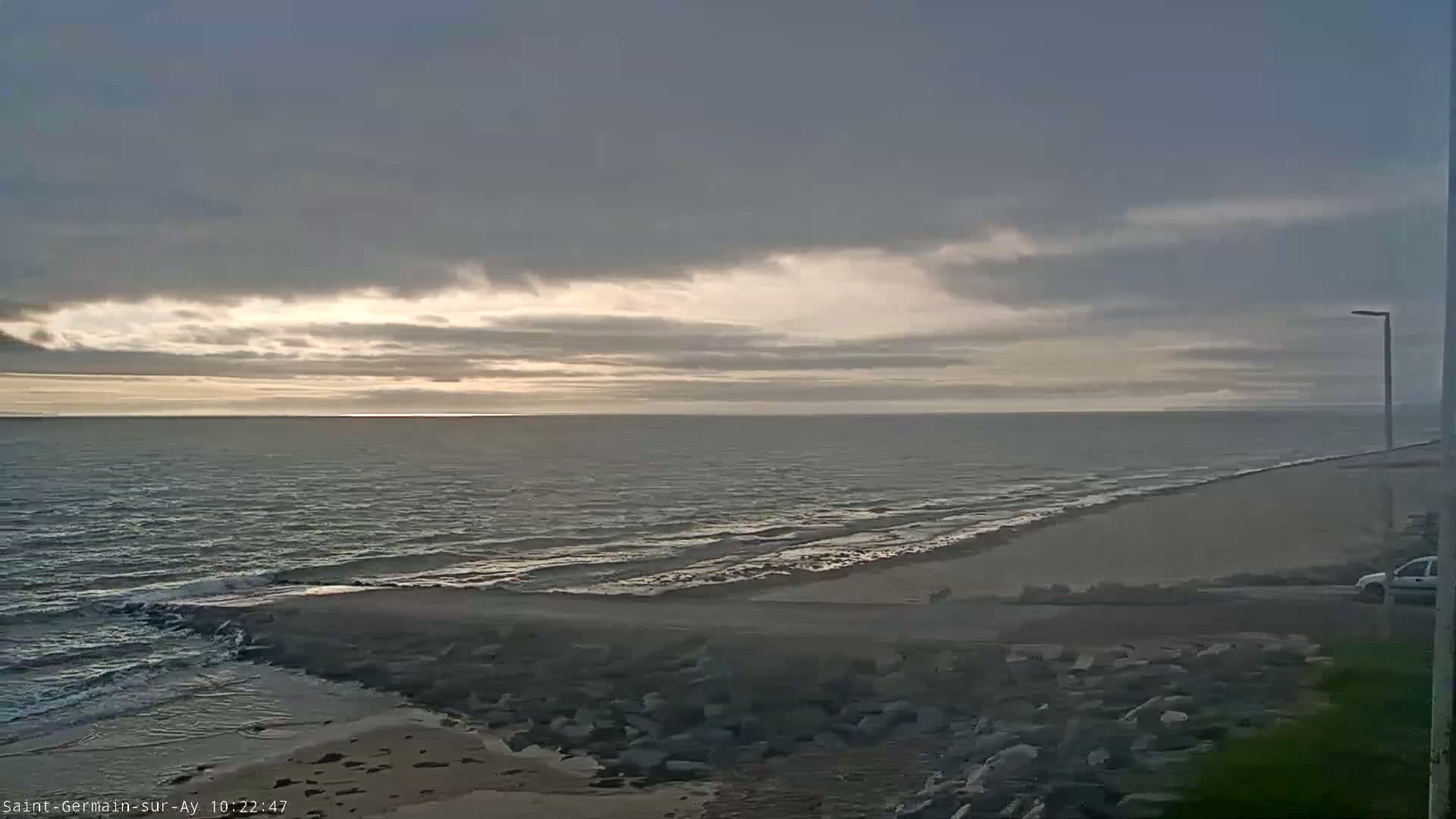 A partly cloudy sky over a calm sea meets a sandy shore with a rocky embankment.