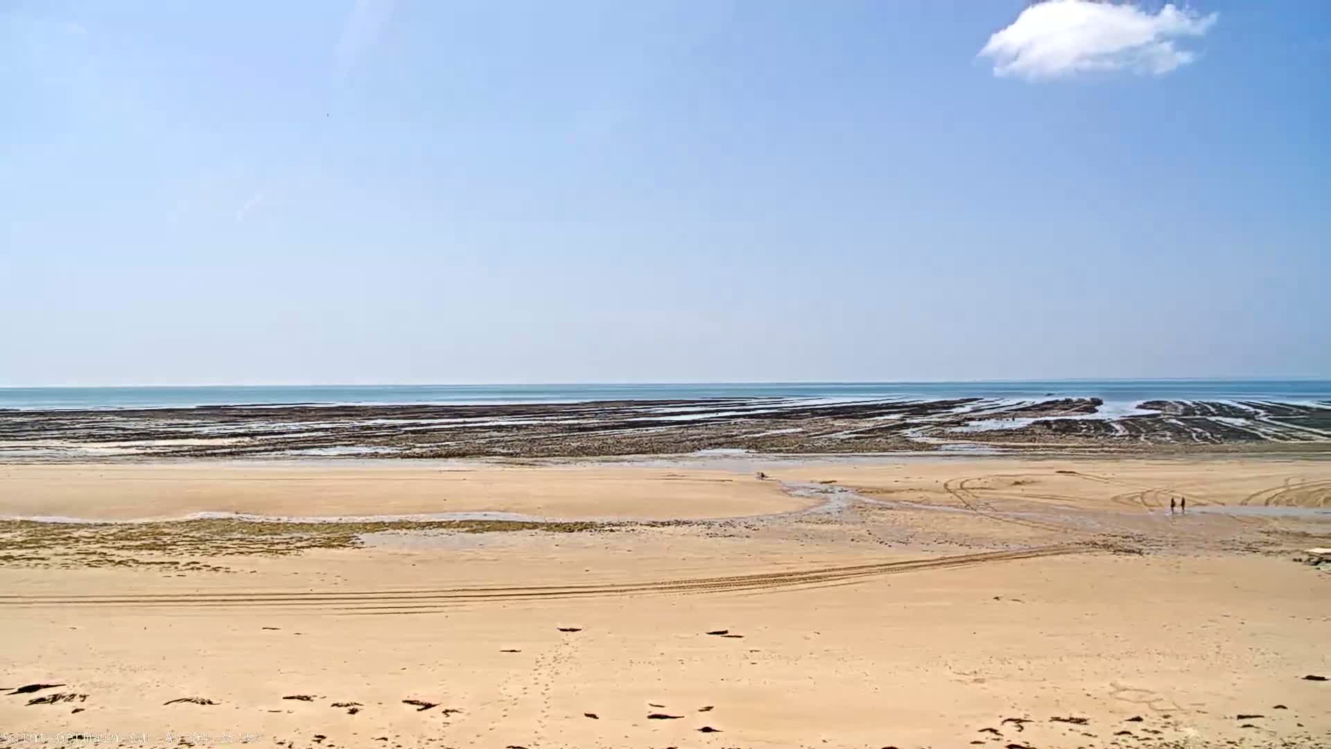 A sandy beach with tire tracks stretches to a calm ocean with dark, layered rock formations visible at low tide under a mostly clear sky with a single small cloud.