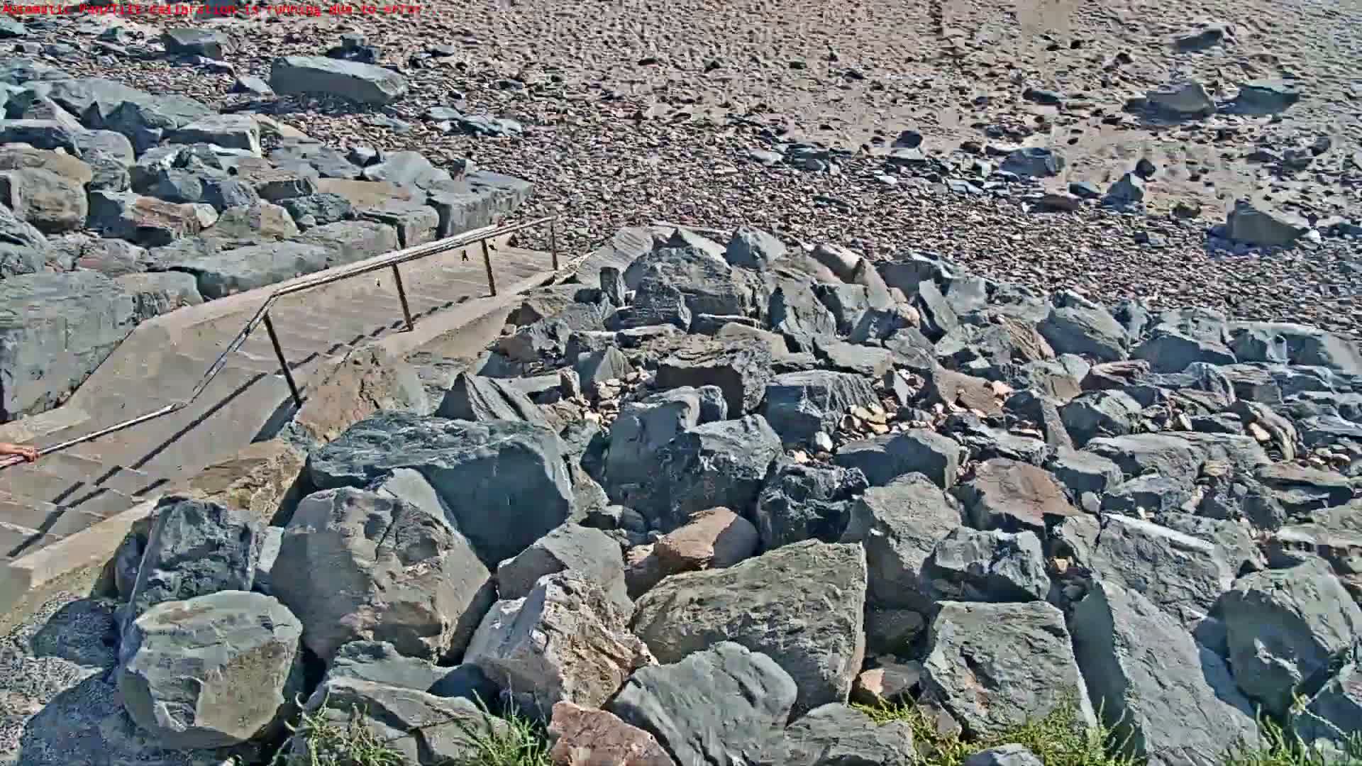 A concrete staircase with a metal railing descends to a rocky beach on a sunny day.