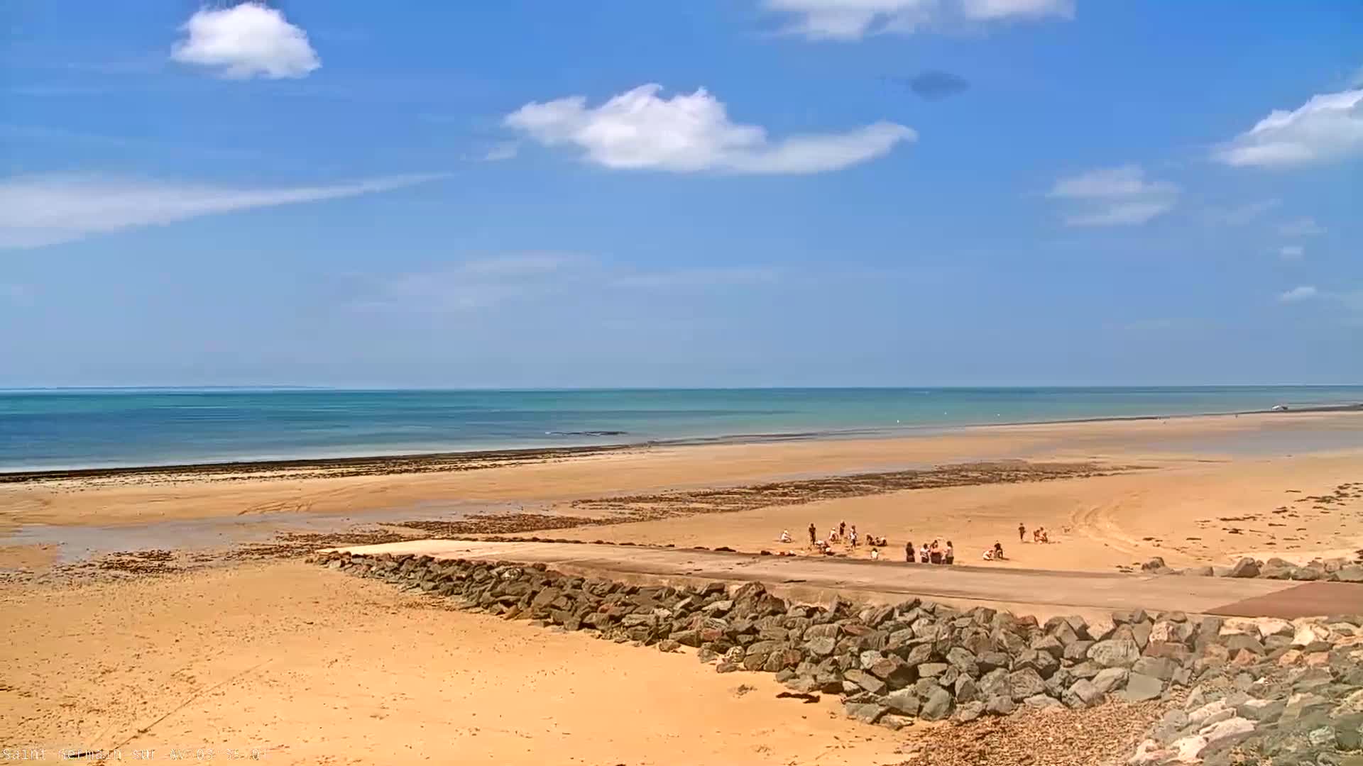 A wide sandy beach stretches to a calm, light-blue ocean under a mostly sunny sky with scattered cumulus clouds.