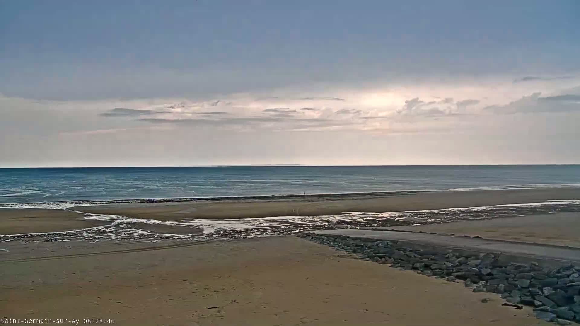 A sandy beach stretches toward a calm, blue ocean under a mostly cloudy sky.