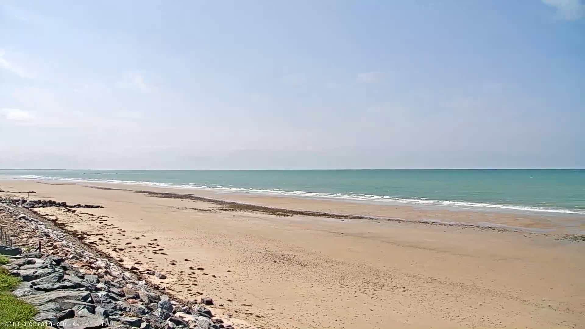 A sandy beach stretches to a calm, light-green ocean under a mostly clear, light-blue sky.