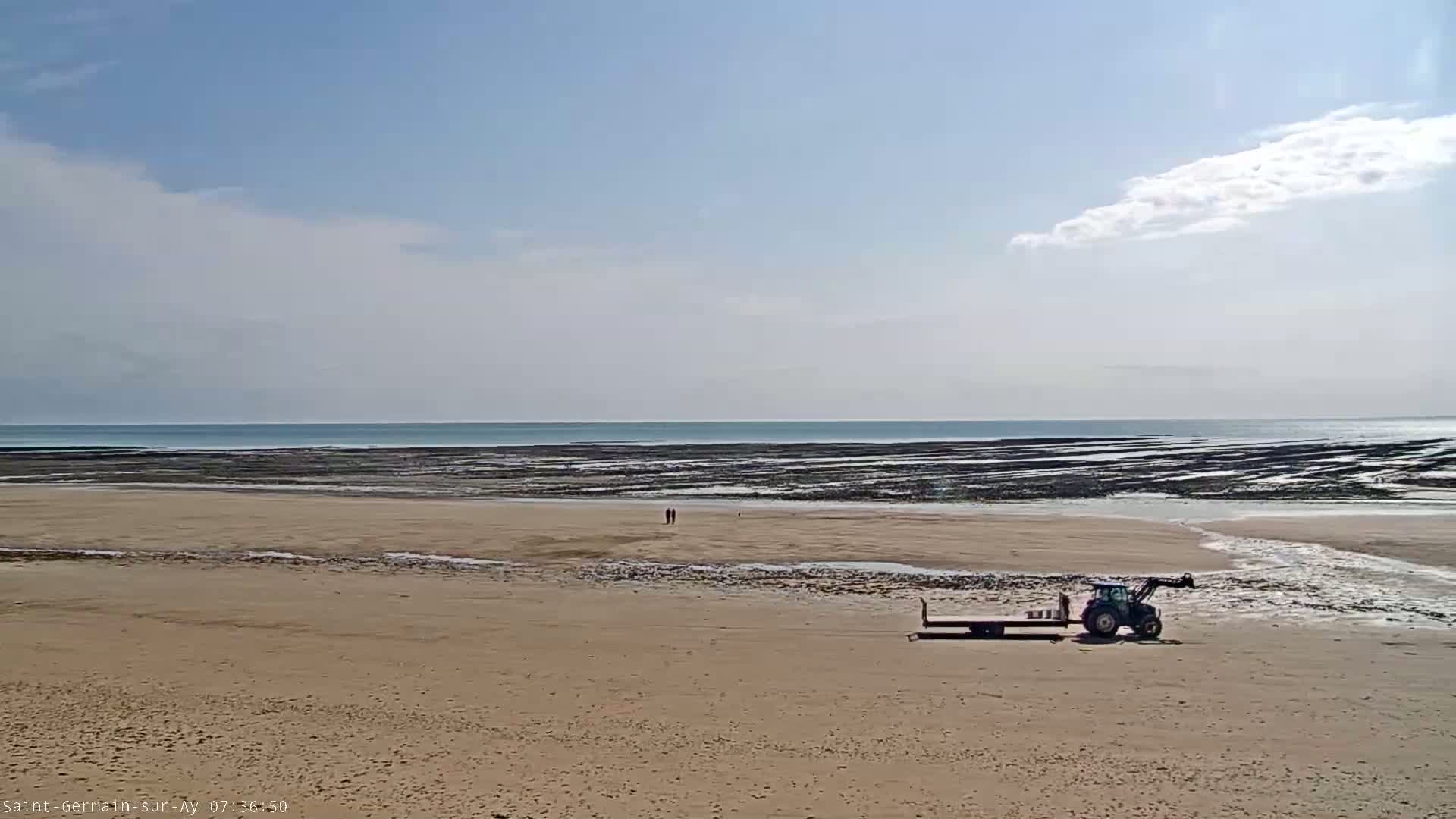 A tractor and trailer sit on a wide sandy beach under a mostly clear sky, with a calm ocean and exposed tidal flats in the background.