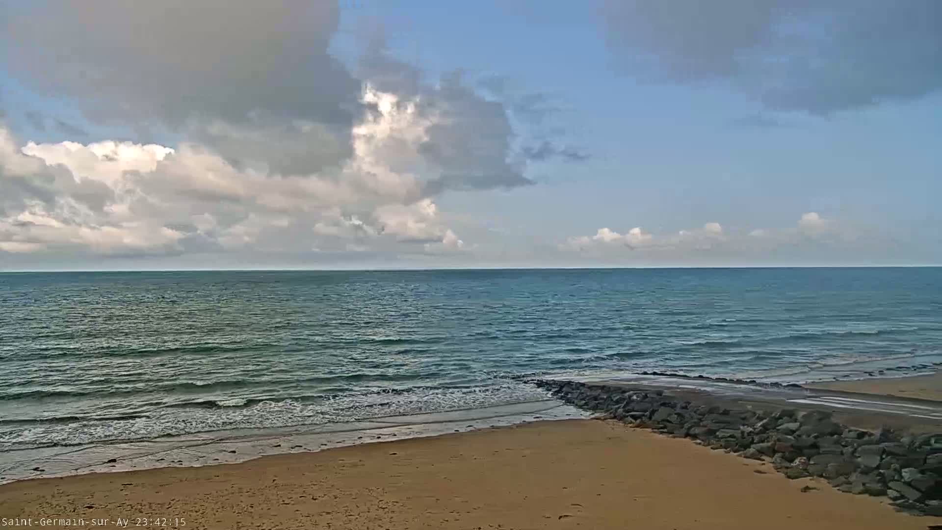 A partly cloudy day shows a sandy beach meeting a calm ocean with small waves at a rocky breakwater.