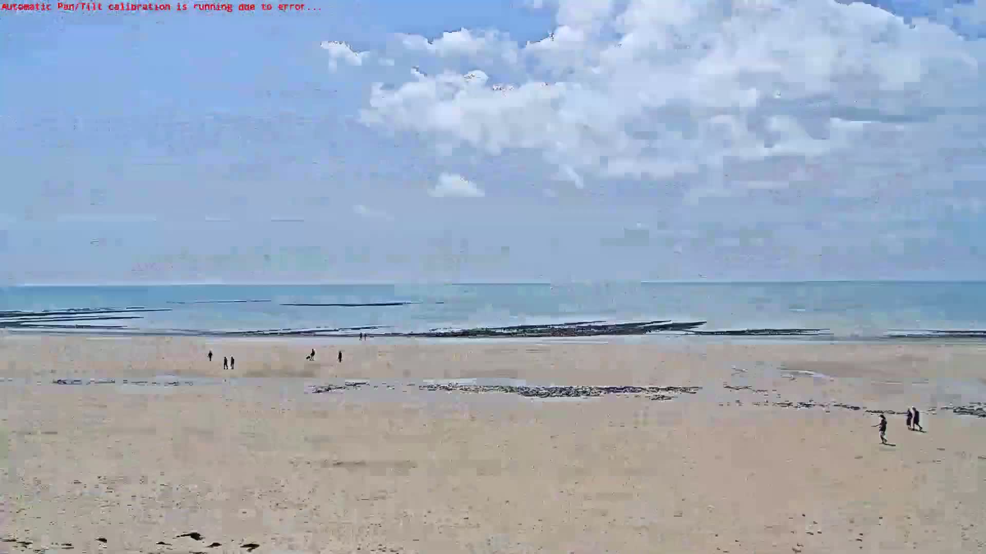 A mostly sunny day shows a wide sandy beach with a few people walking, leading to a calm ocean under a partly cloudy sky.