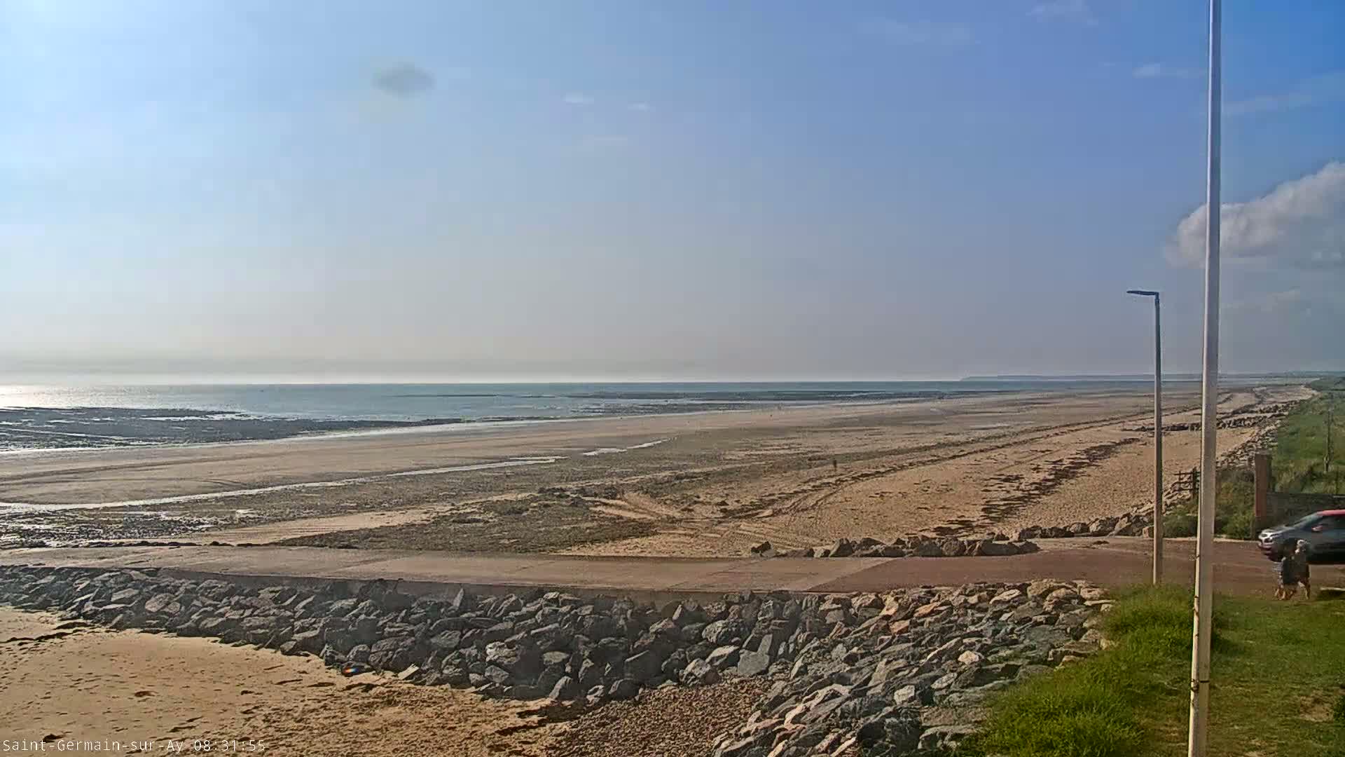 A wide sandy beach stretches to a calm ocean under a mostly clear, sunny sky.
