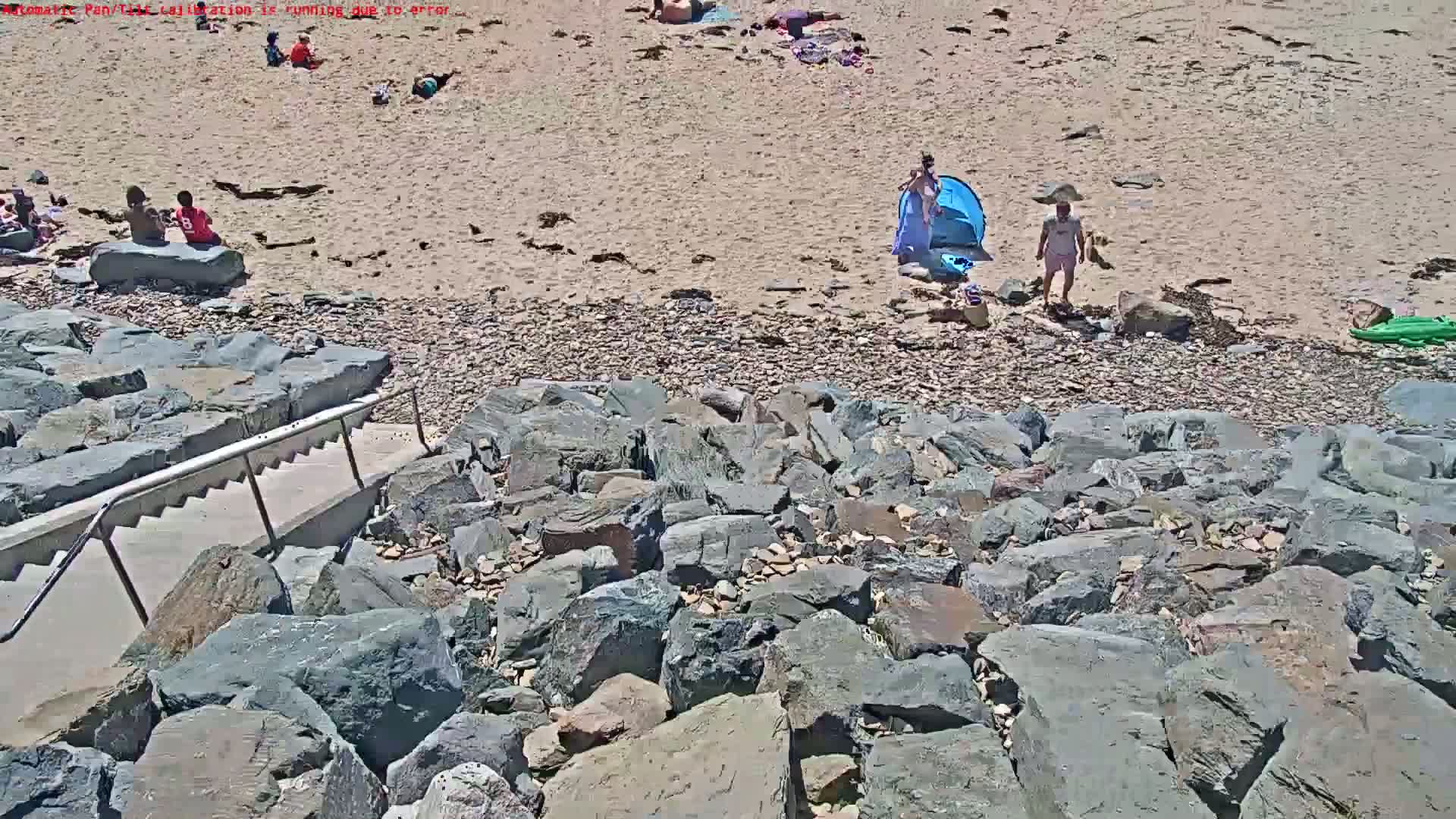 A rocky staircase leads down to a sandy beach where several people are relaxing on a sunny day.