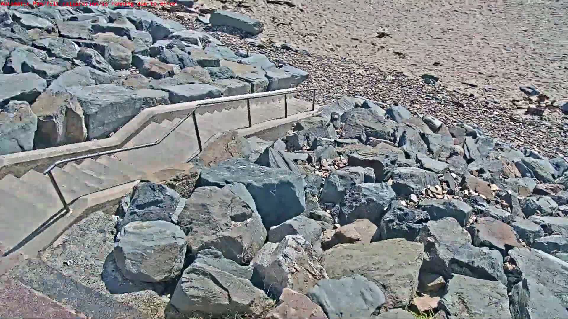 A concrete staircase with metal railings descends to a sandy beach bordered by large, dark gray rocks under sunny conditions.