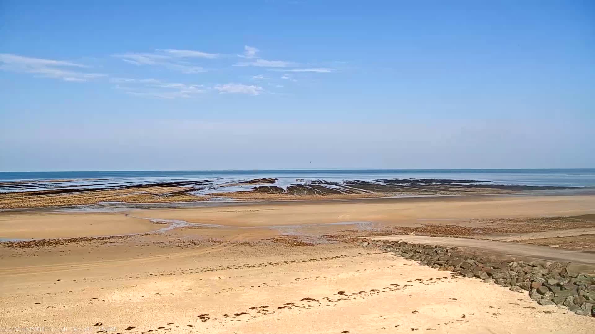 A sandy beach stretches to a calm ocean under a clear blue sky with a few scattered clouds.