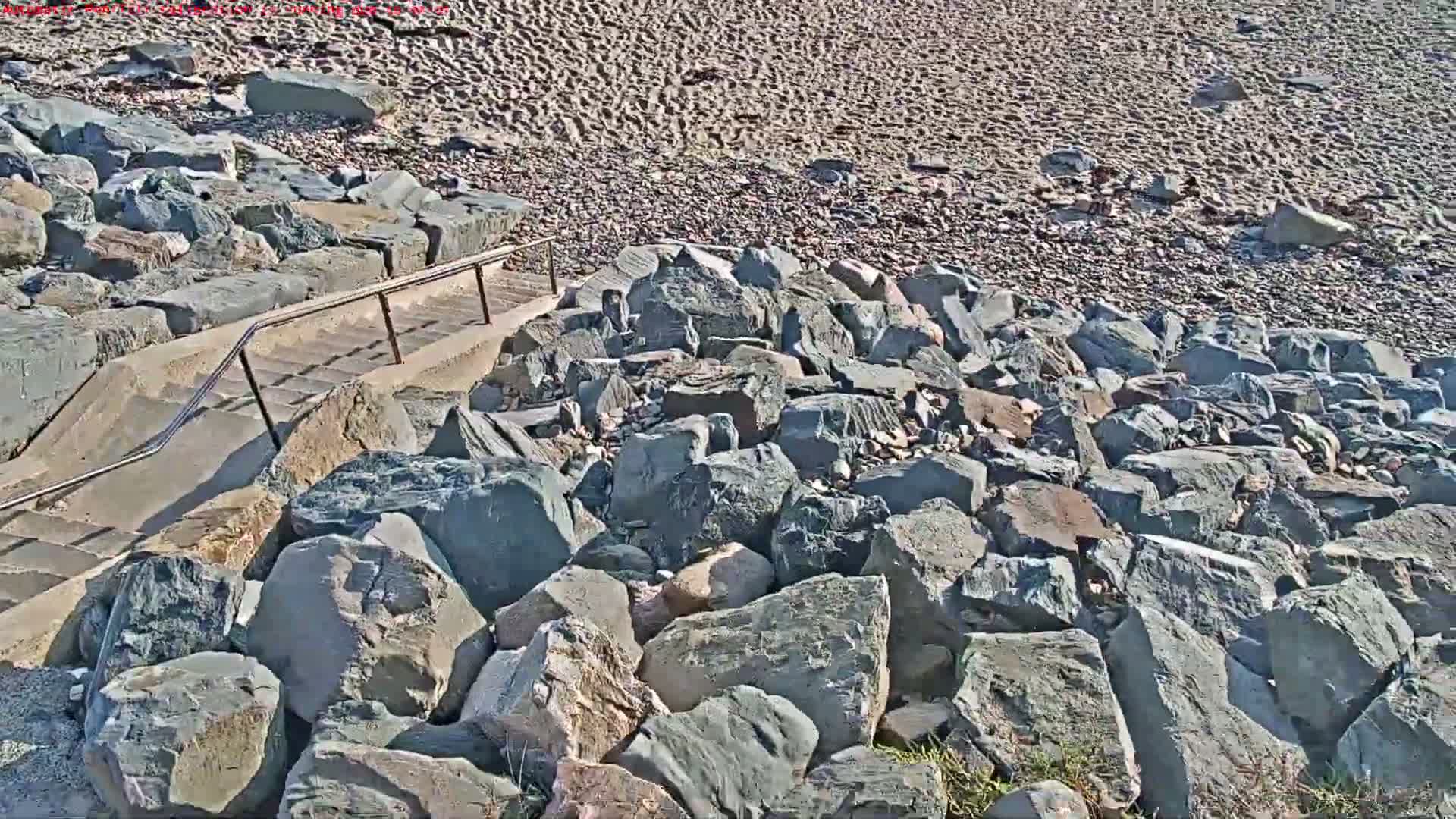 A concrete staircase with a metal railing leads down to a rocky beach with a sandy area visible in the background.