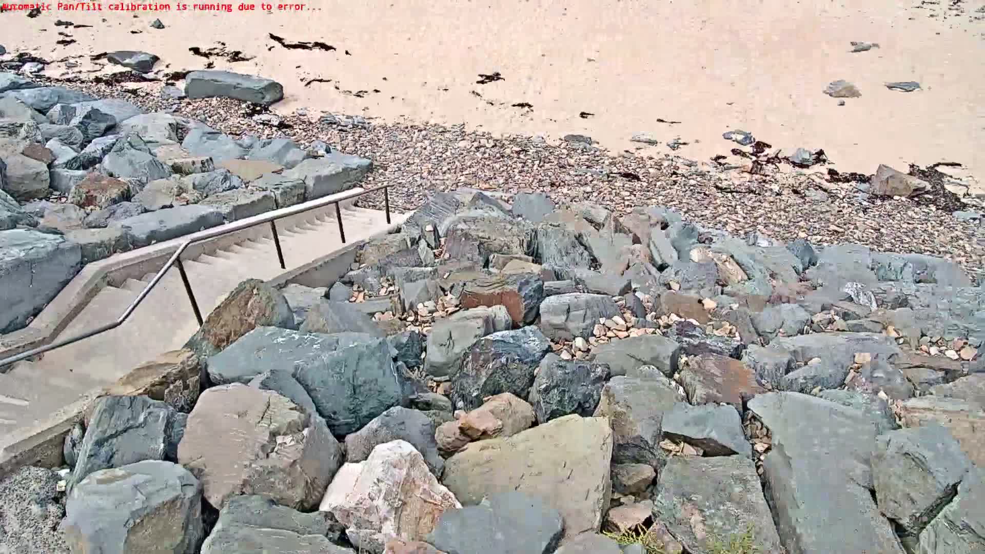 A concrete staircase with a metal railing descends to a rocky beach with light-colored sand.