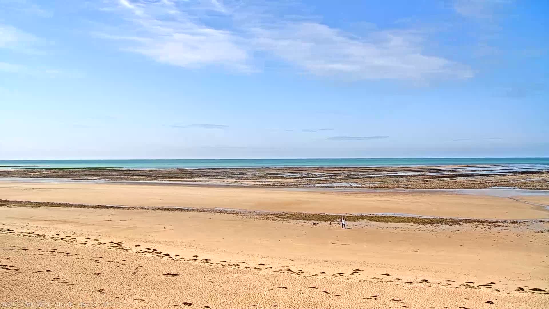 A wide sandy beach stretches to a calm, light turquoise ocean under a mostly sunny sky with a few scattered clouds.