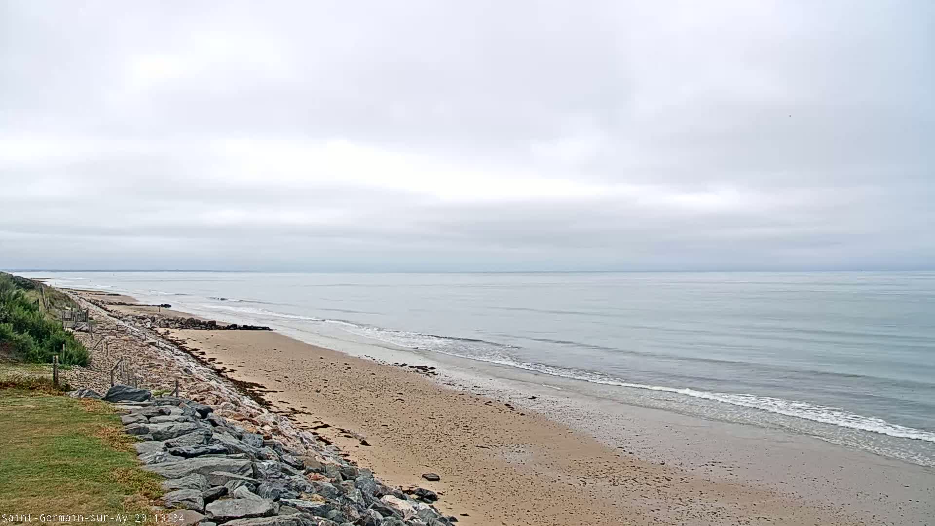 A sandy beach with gentle waves stretches along a calm, overcast ocean.