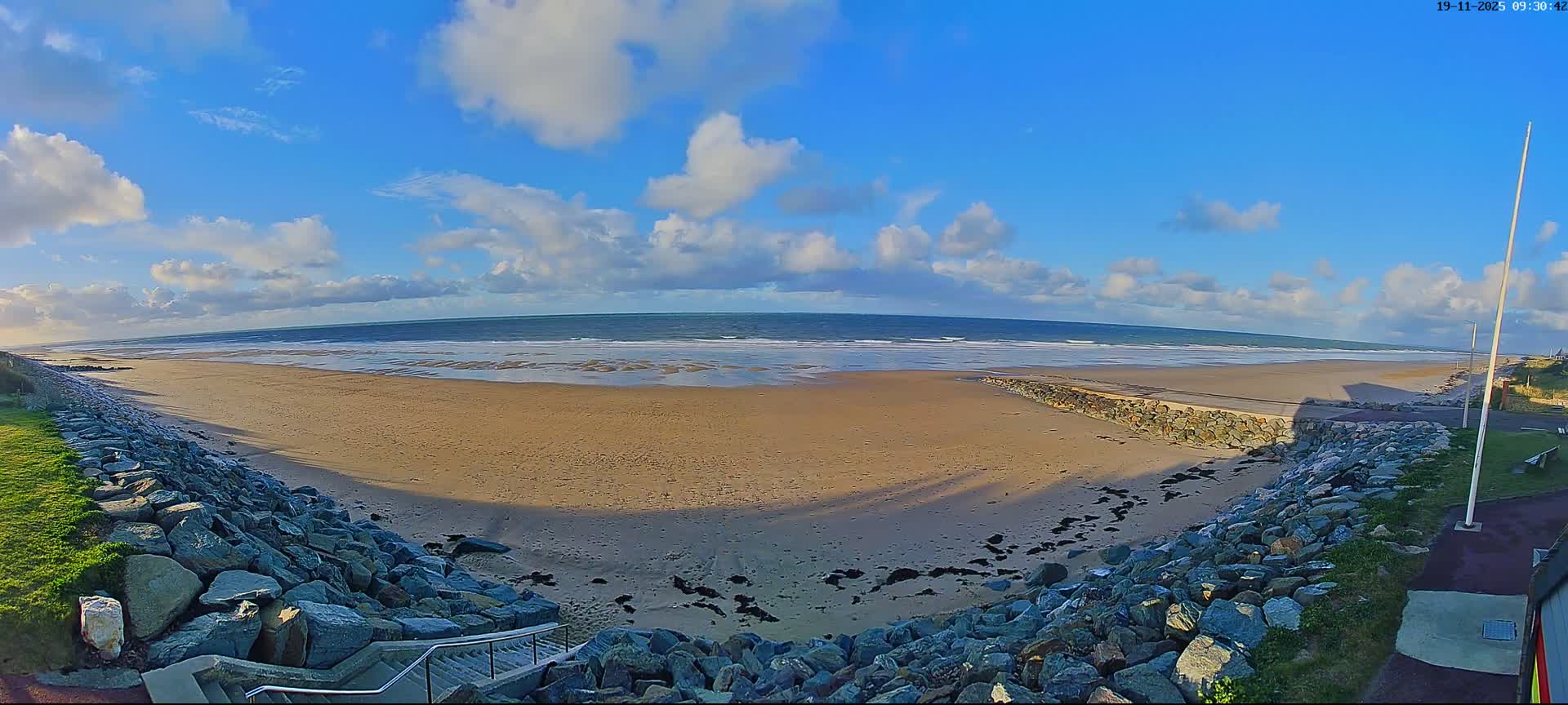 A wide-angle view captures a sunny beach with scattered white clouds in a bright blue sky, featuring a vast sandy expanse, gentle ocean waves, and rocky sea defenses lining the foreground.