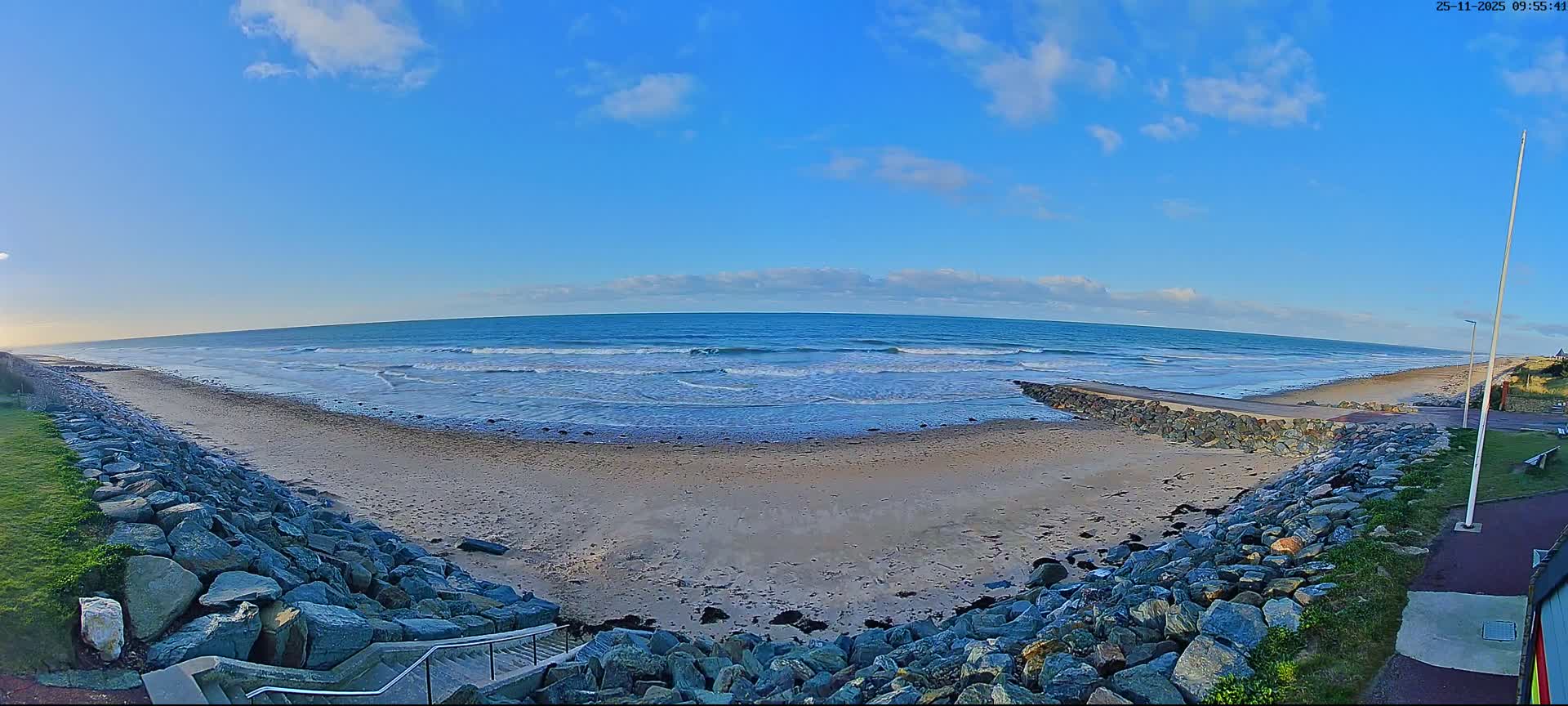A panoramic view reveals a wide sandy beach with breaking waves under a bright blue sky with scattered clouds, flanked by rock seawalls on both sides leading to a set of stairs.