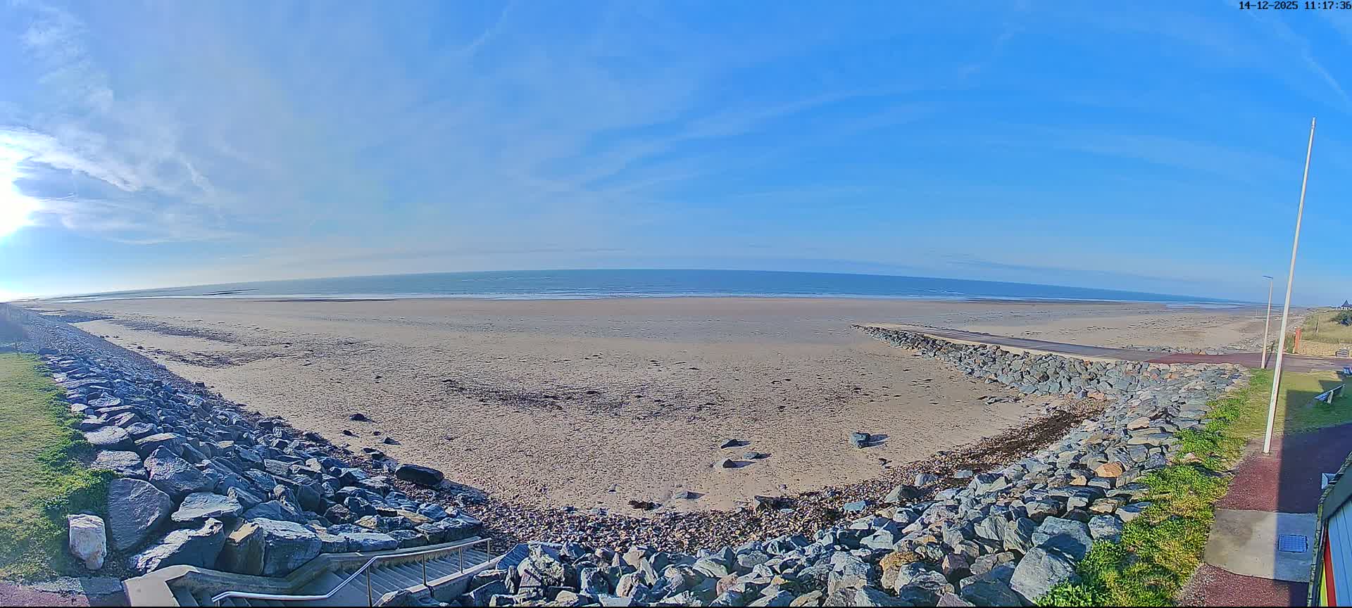 A panoramic view reveals a wide sandy beach with breaking waves under a bright blue sky with scattered clouds, flanked by rock seawalls on both sides leading to a set of stairs.