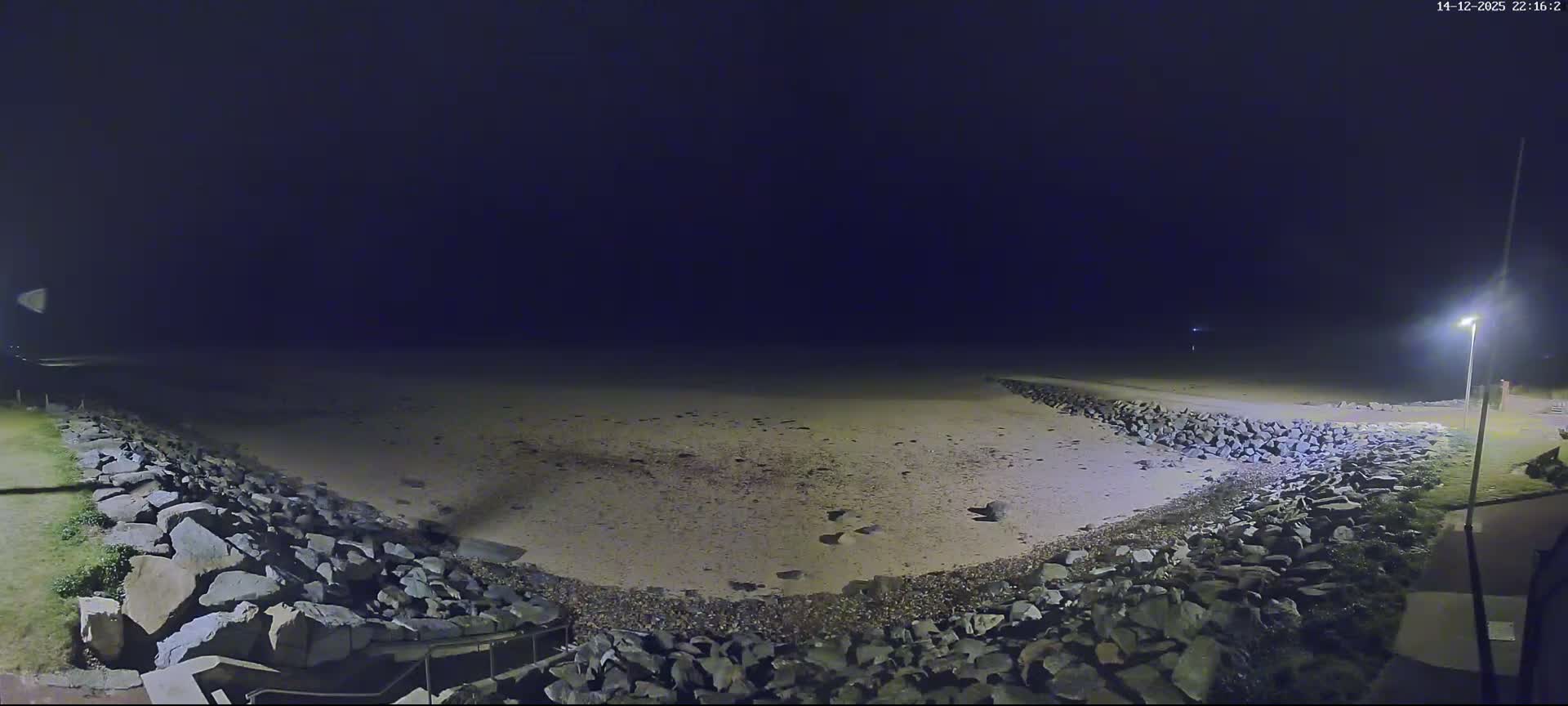 A panoramic view reveals a wide sandy beach with breaking waves under a bright blue sky with scattered clouds, flanked by rock seawalls on both sides leading to a set of stairs.