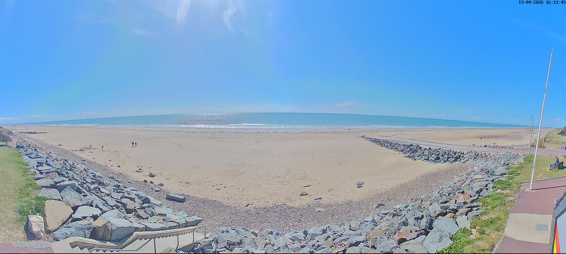 A panoramic view reveals a wide sandy beach with breaking waves under a bright blue sky with scattered clouds, flanked by rock seawalls on both sides leading to a set of stairs.