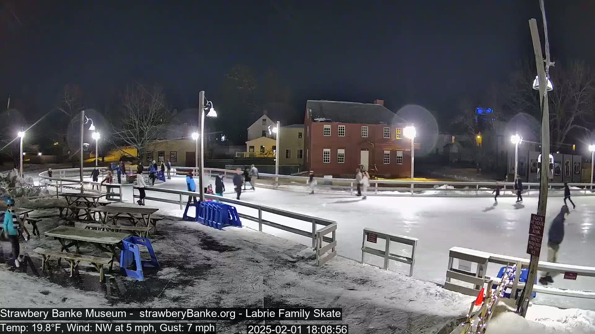 People ice skate at night on an outdoor rink illuminated by several lights, with snow on the ground and a nearby building visible.