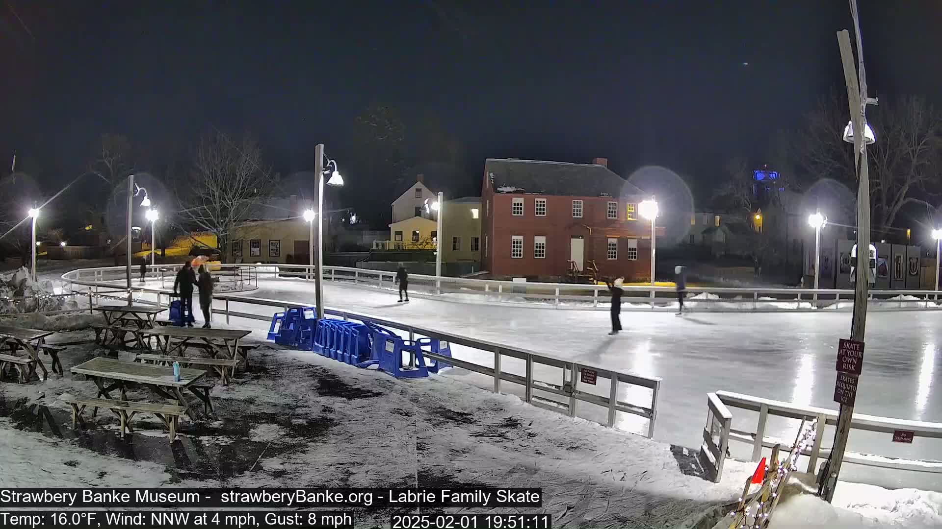 At night, several people ice skate on an outdoor rink lit by lampposts, with picnic tables and snow nearby under a dark sky.