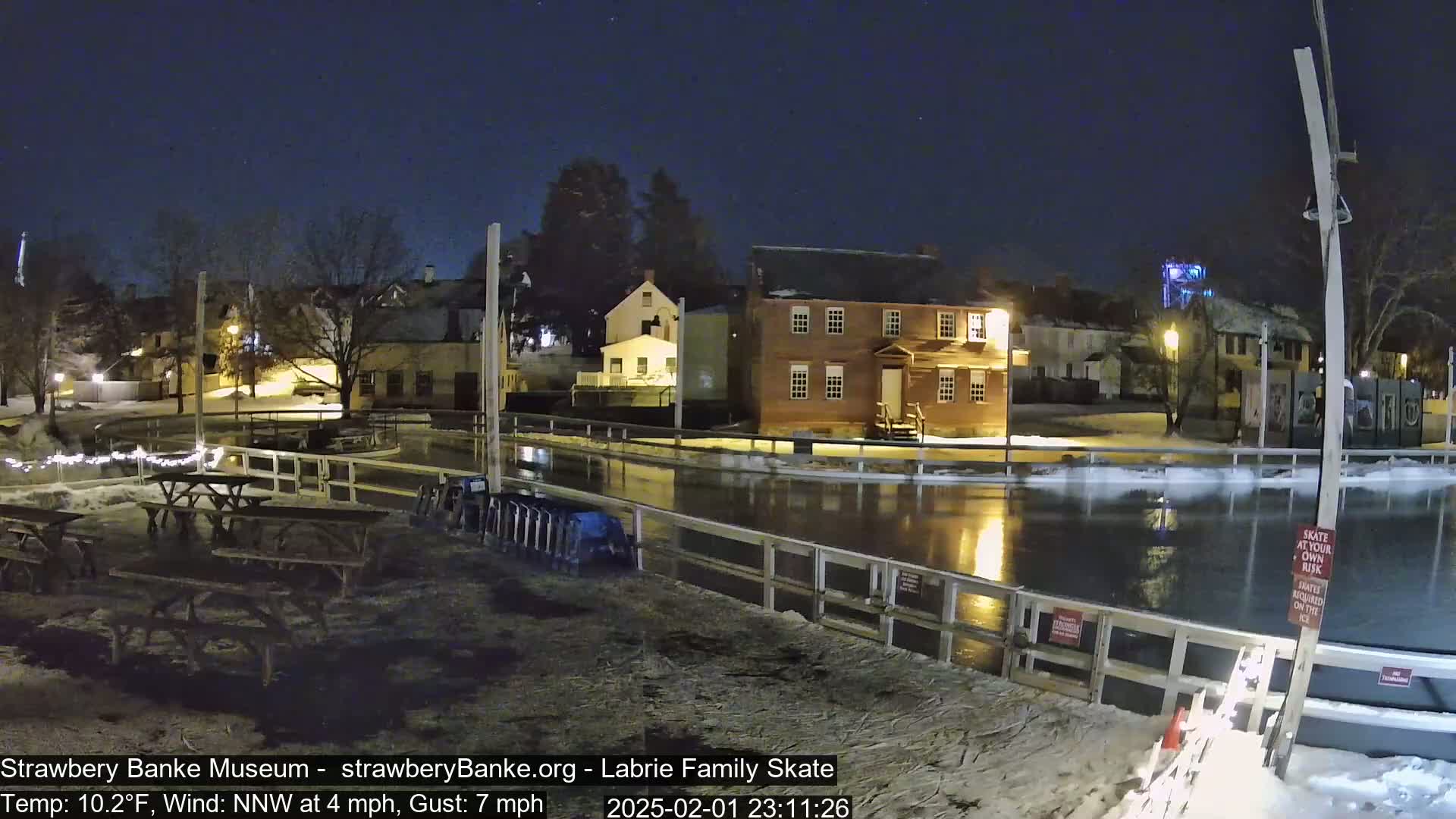 A nighttime view of a partially frozen outdoor ice rink surrounded by historic buildings under a clear, dark sky.