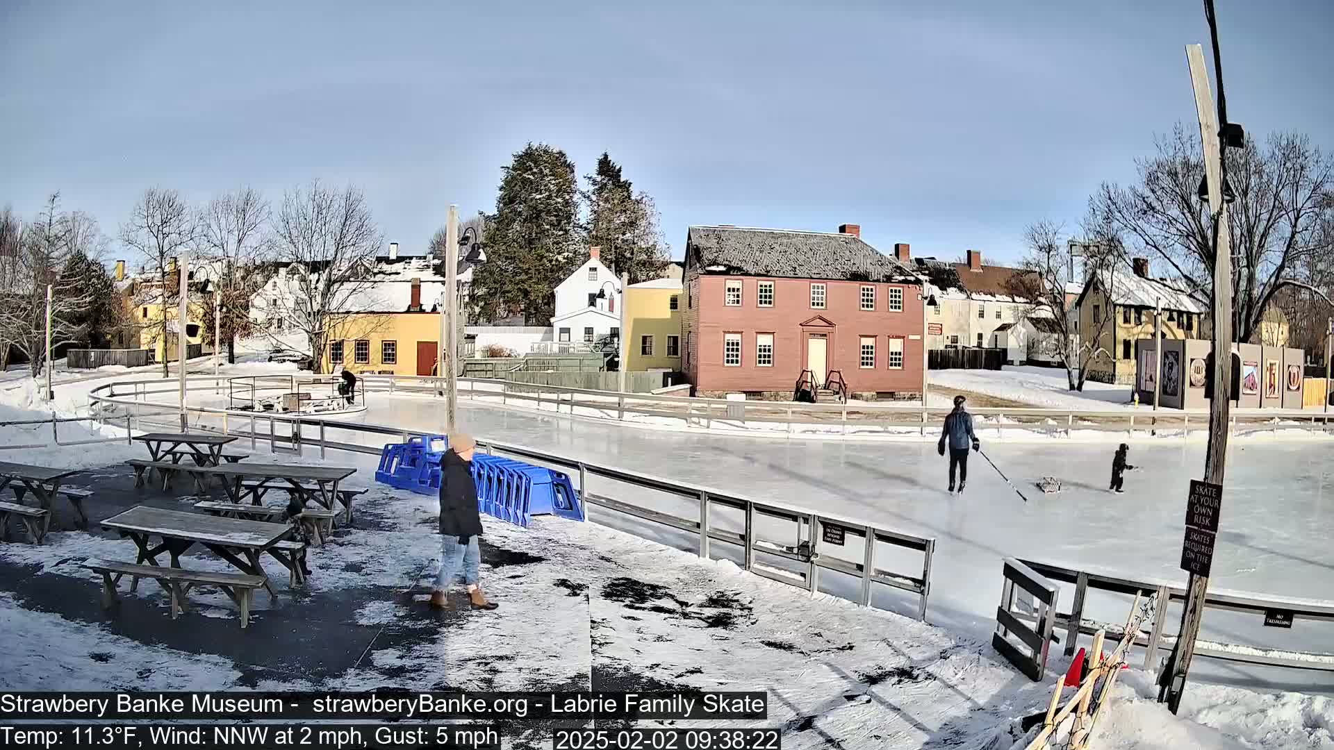 An outdoor ice skating rink, surrounded by snow-covered ground and historic-looking buildings, is partially occupied by several people skating and others standing near picnic tables on a sunny, cold day.