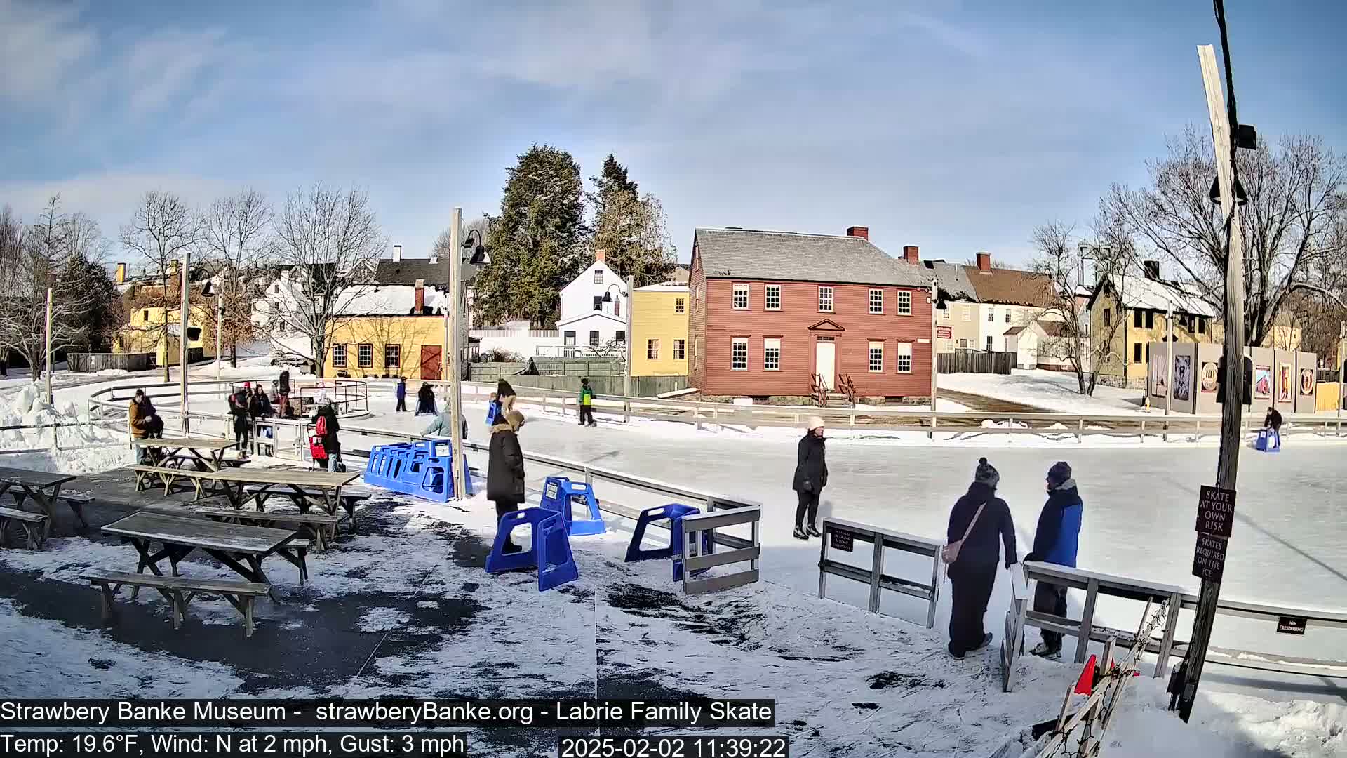 An outdoor ice-skating rink, surrounded by snow-covered ground and historic-looking buildings, is under a sunny sky.