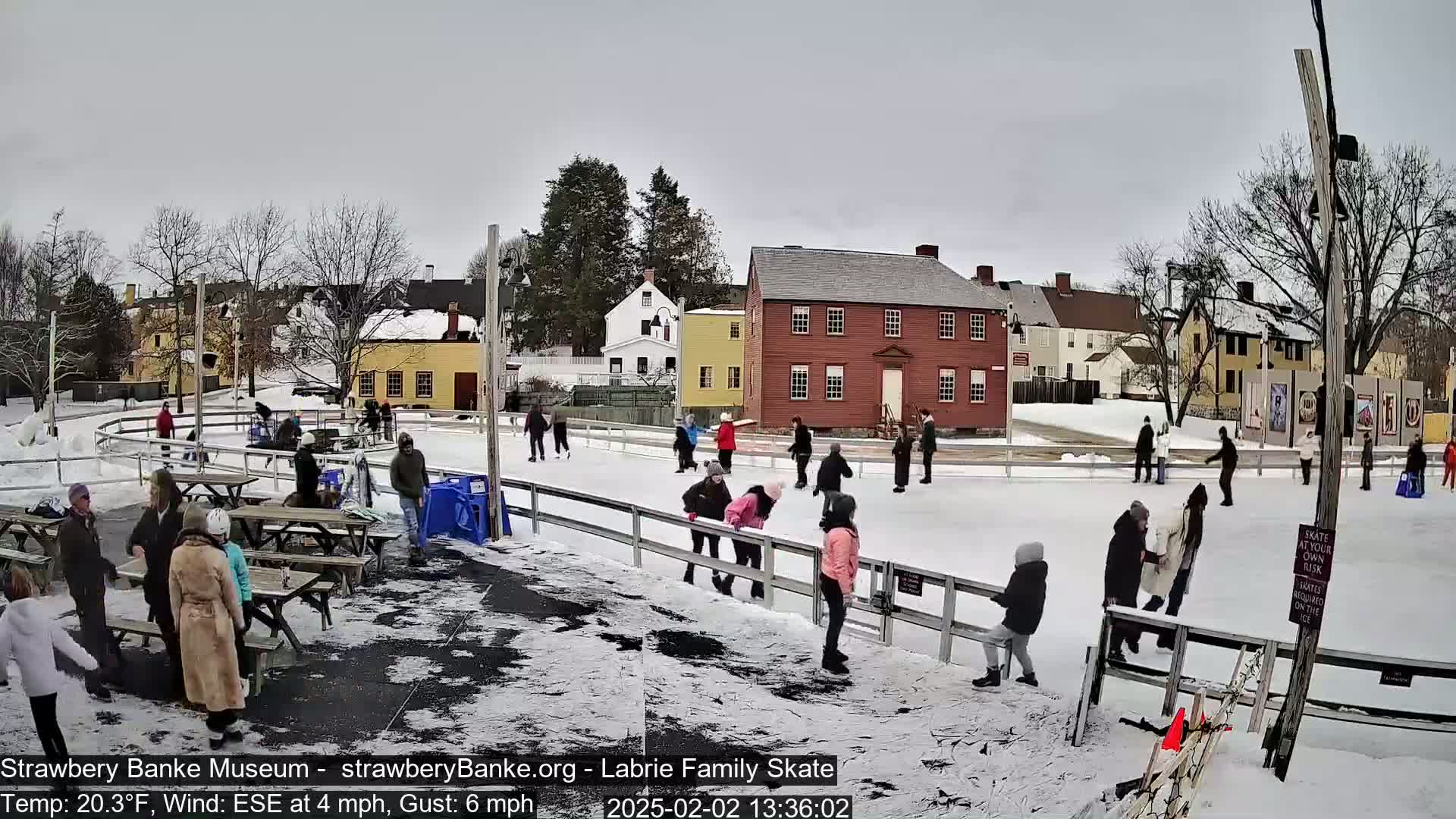 An outdoor ice skating rink, surrounded by snow-covered ground and historical-looking buildings, is populated by several ice skaters on a lightly overcast day.