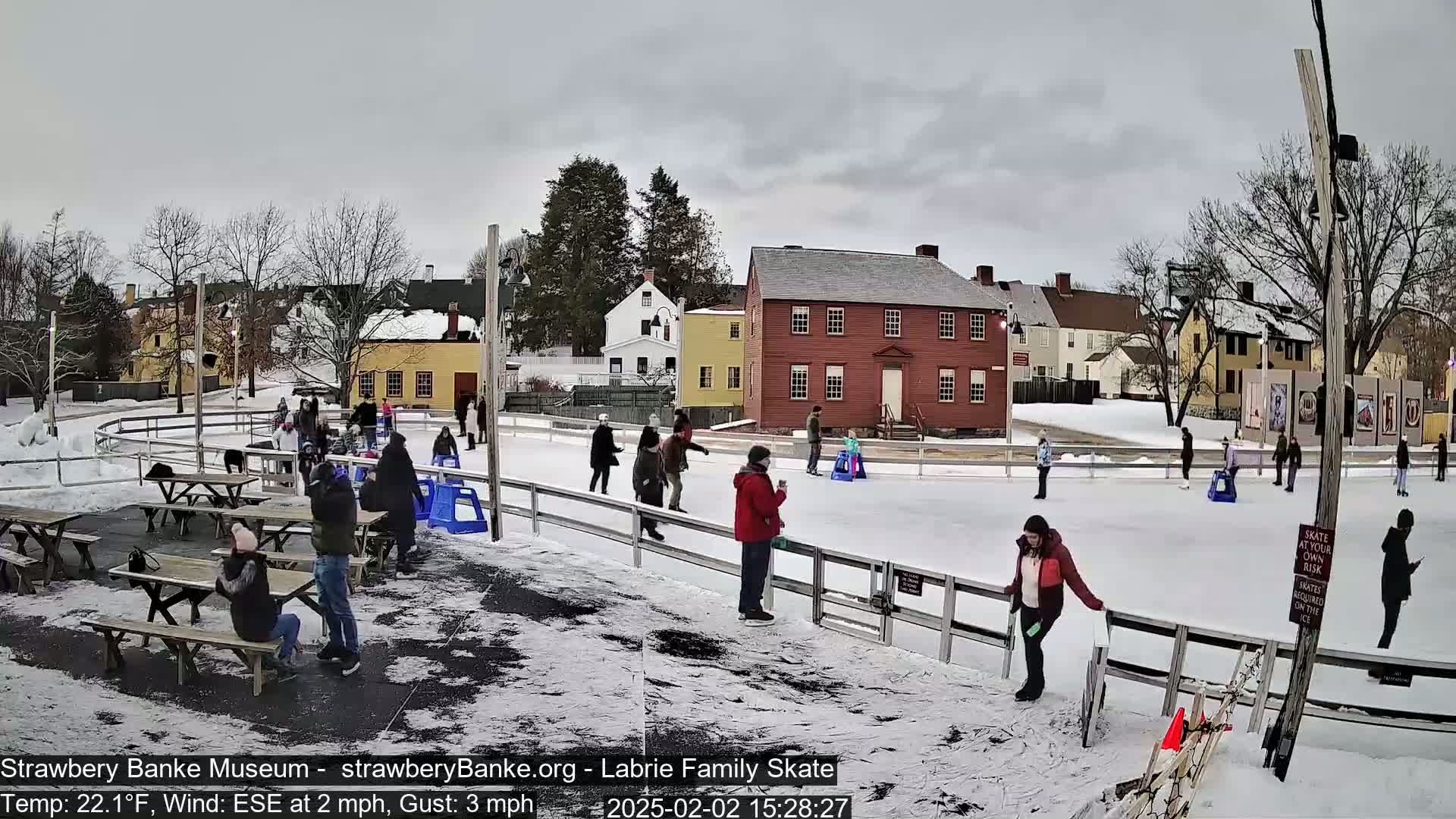 People ice skate on an outdoor rink surrounded by snow-covered ground and historic-looking buildings under an overcast sky.