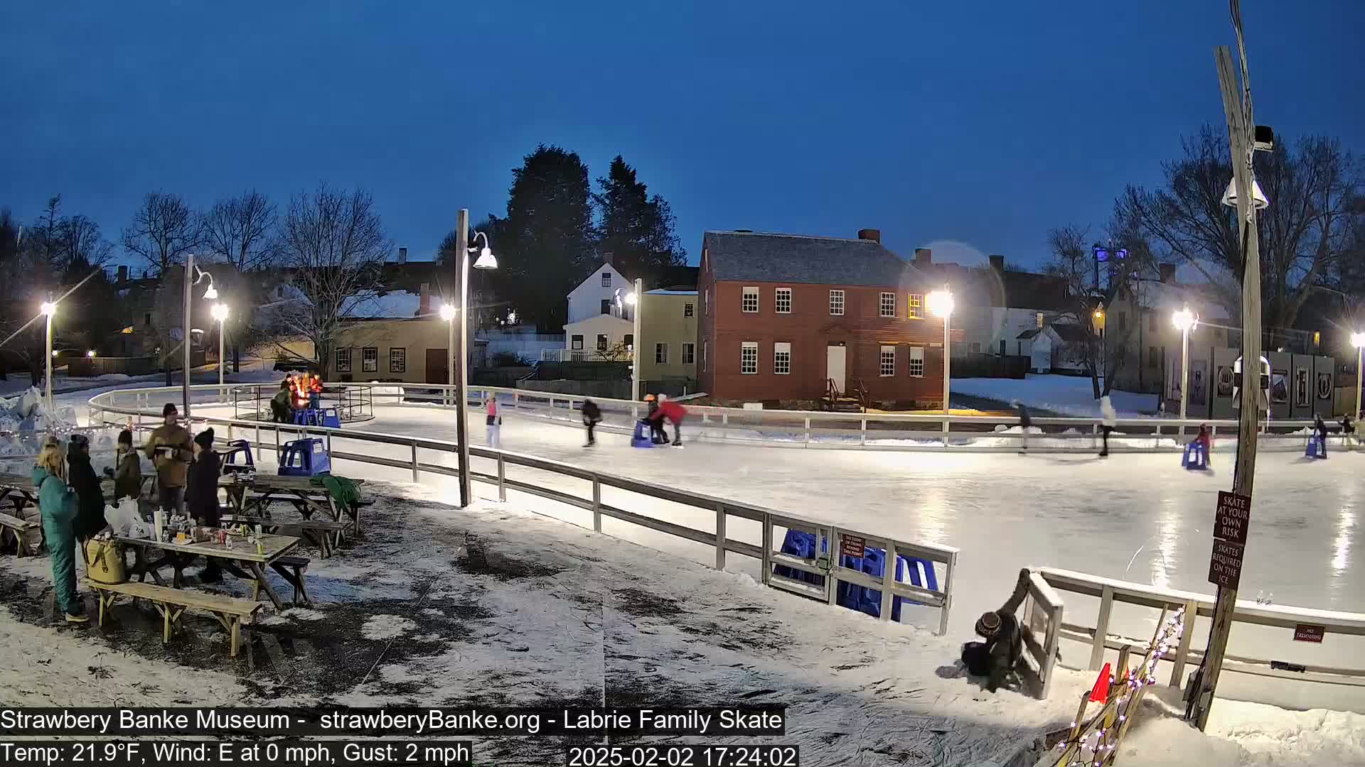 An outdoor ice skating rink at night, illuminated by lampposts, with several people ice skating and others gathered at picnic tables nearby, under a clear, dark blue twilight sky.