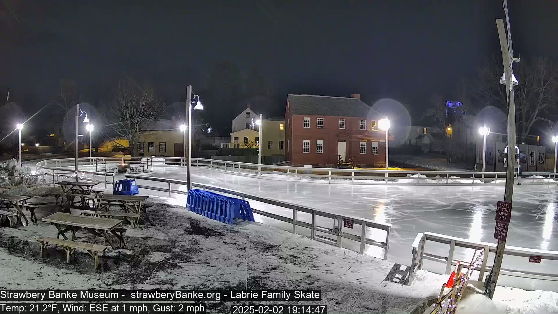 An outdoor ice-skating rink is lit at night by several lampposts, surrounded by snow and picnic tables near some historic-looking buildings.