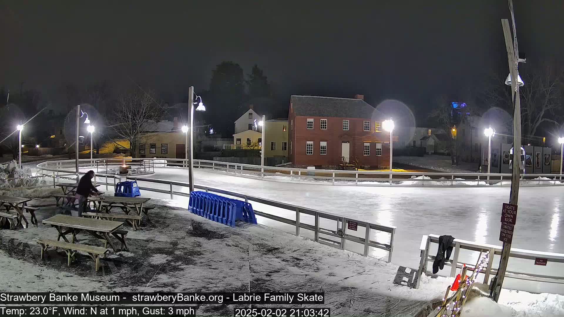 A nighttime view of an outdoor ice rink illuminated by several lights, with a few people near picnic tables and a building in the background, under light snowfall.