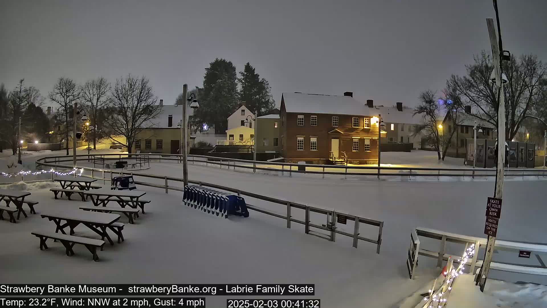 A snow-covered outdoor ice rink at night, surrounded by snow-covered picnic tables and historic buildings, under a dark, overcast sky.